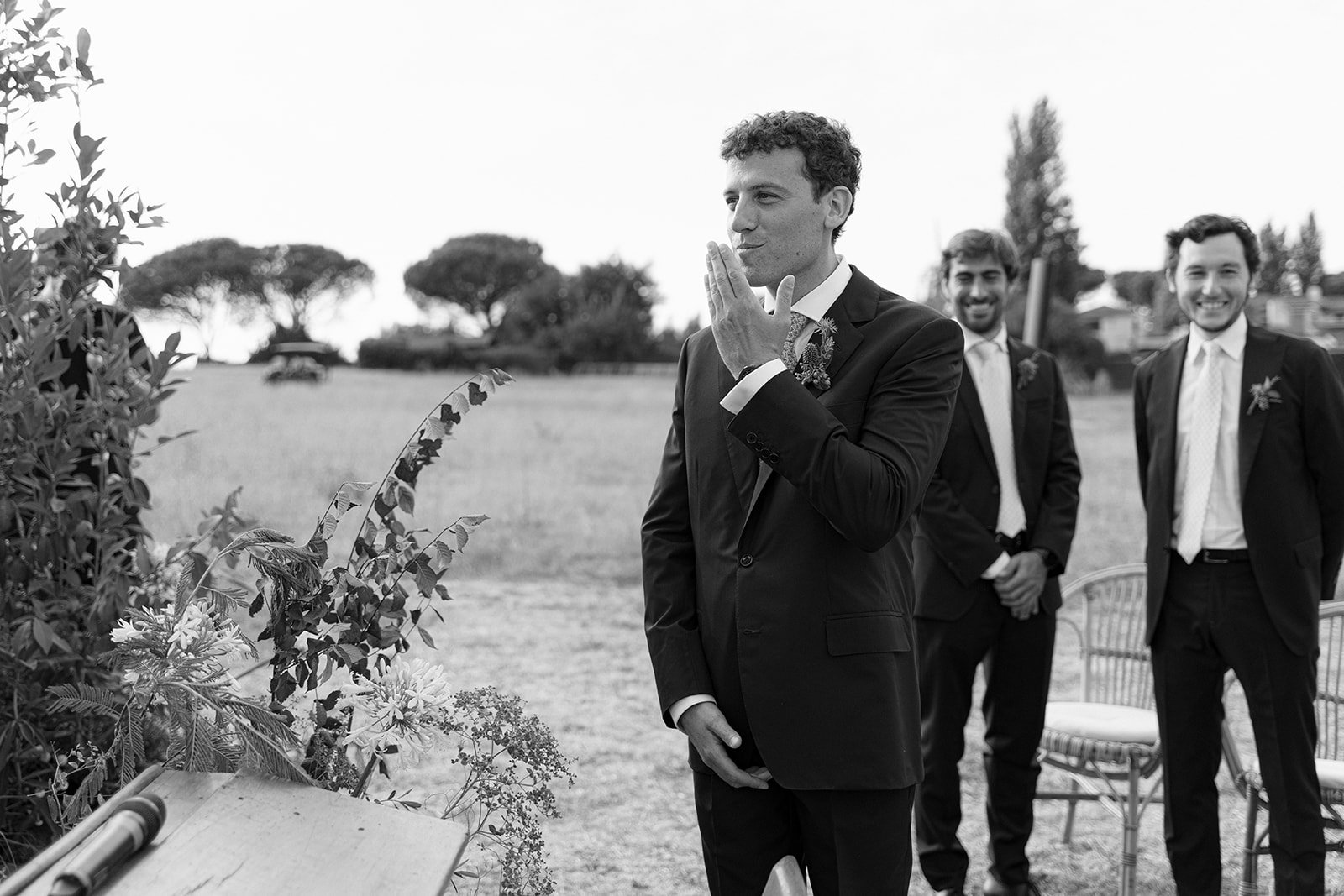 Black and white image of a groom with curled hair in a suit, touching his lips, standing outdoors with two men in suits smiling in the background, and trees and houses in the distance.