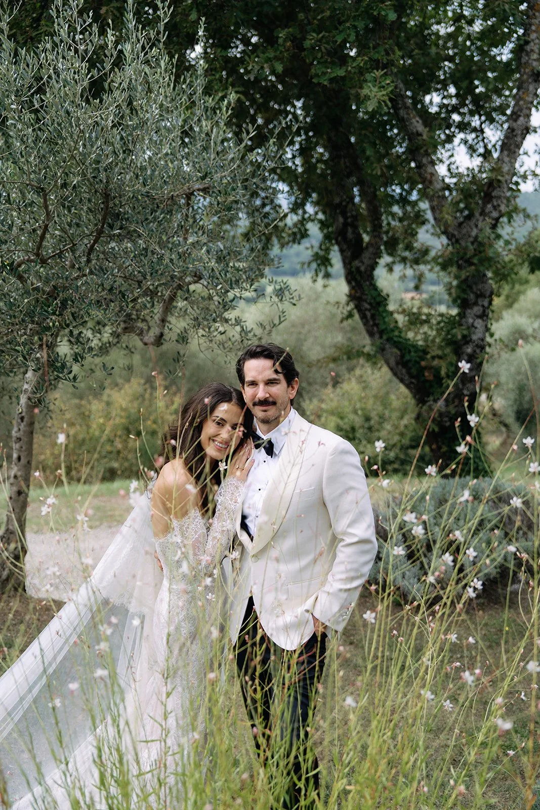 A bride and groom standing among tall grass and wildflowers outdoors, smiling at the camera with trees and greenery in the background.