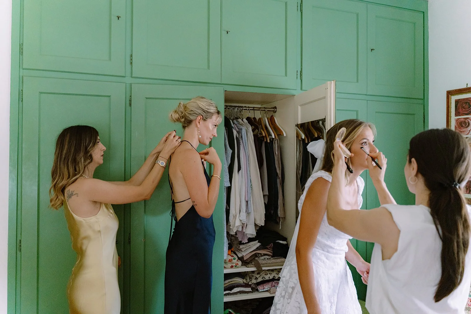 Four women in a bedroom with green cabinets, one is fixing another's hair, another is applying makeup while the bride prepares.