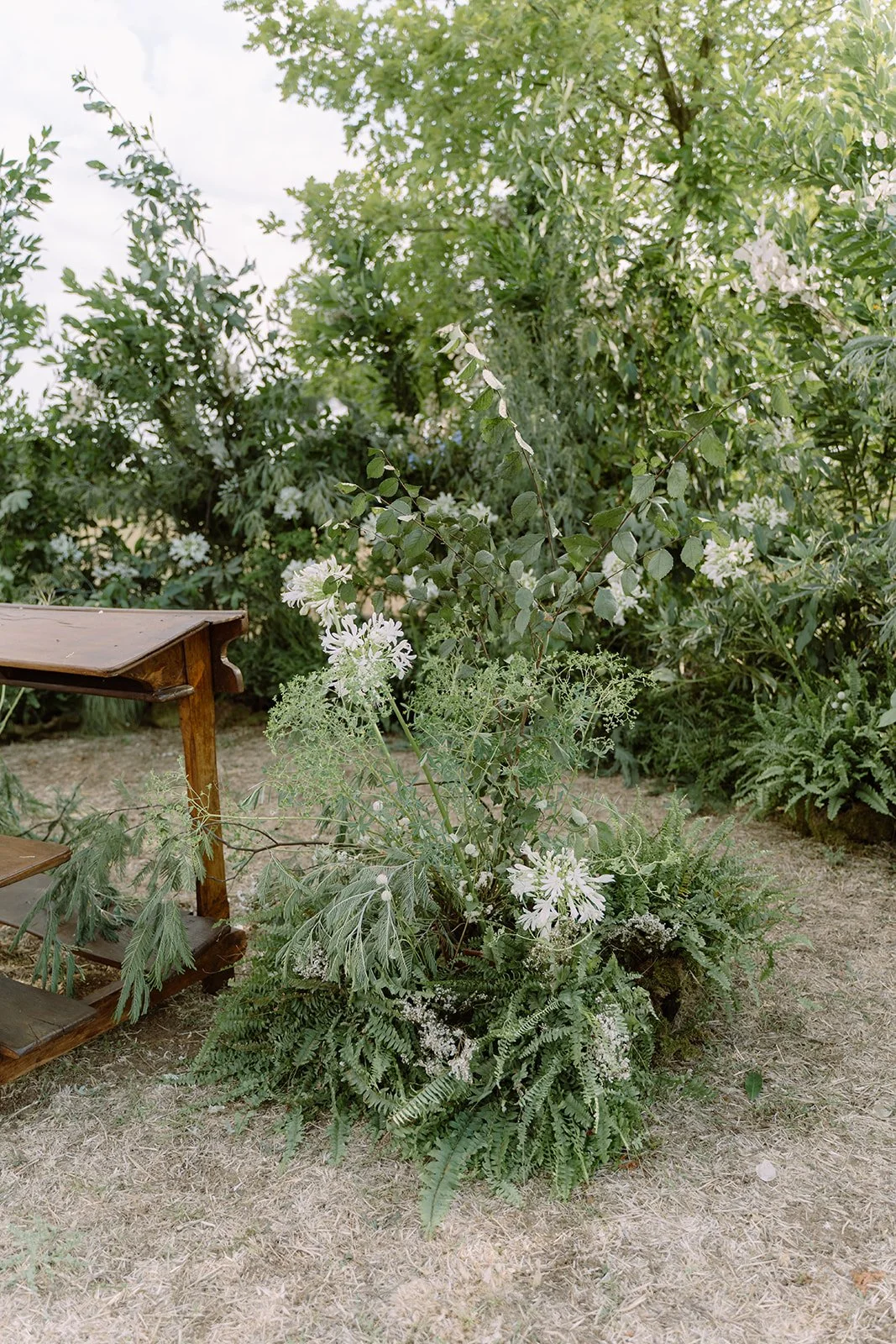A garden scene with a large plant arrangement of white flowers and greenery on the ground, next to a wooden table on the left side, and surrounded by dense green bushes and trees in the background.