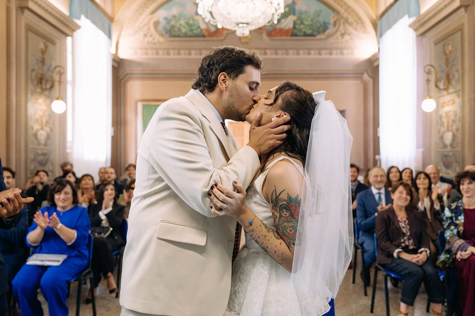 A groom in a cream suit and a bride with a veil kissing during their wedding ceremony in a decorated hall, with guests seated and clapping in the background.