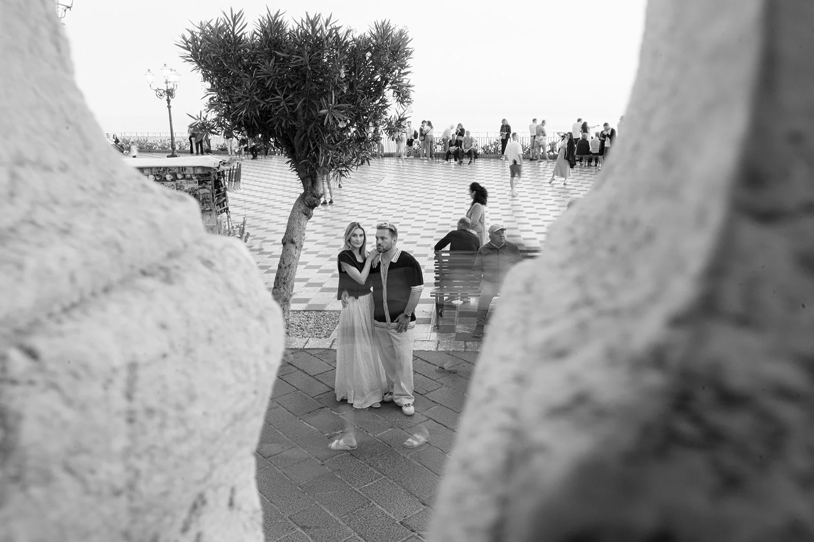 A black-and-white photo taken through a small opening in a stone wall showing a couple posing on a sidewalk near a tree, with a large open square filled with people in the background.