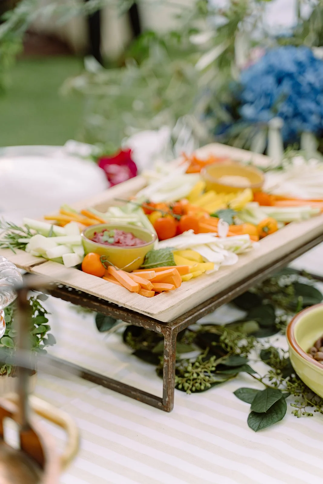 Wooden platter with sliced vegetables including carrots, celery, and cherry tomatoes, accompanied by dipping sauces, on a table with greenery beneath.