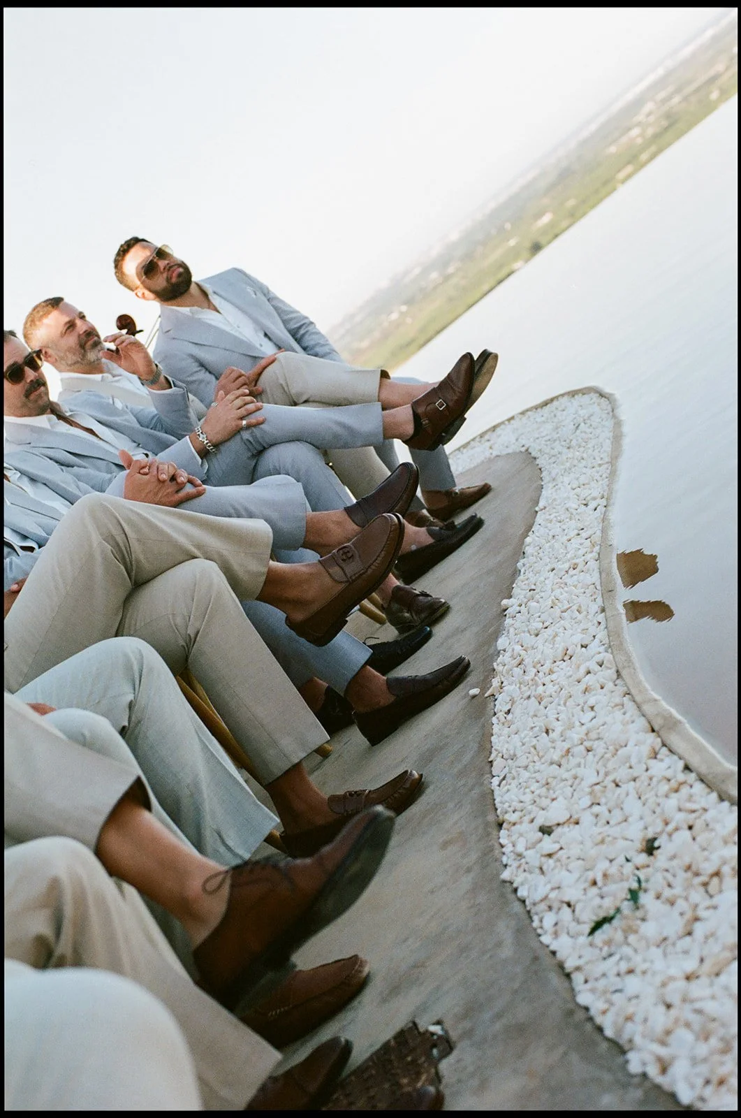 Group of men in suits sitting on chairs by a pond, dressed in light-colored clothing and sunglasses, outdoors during the daytime.