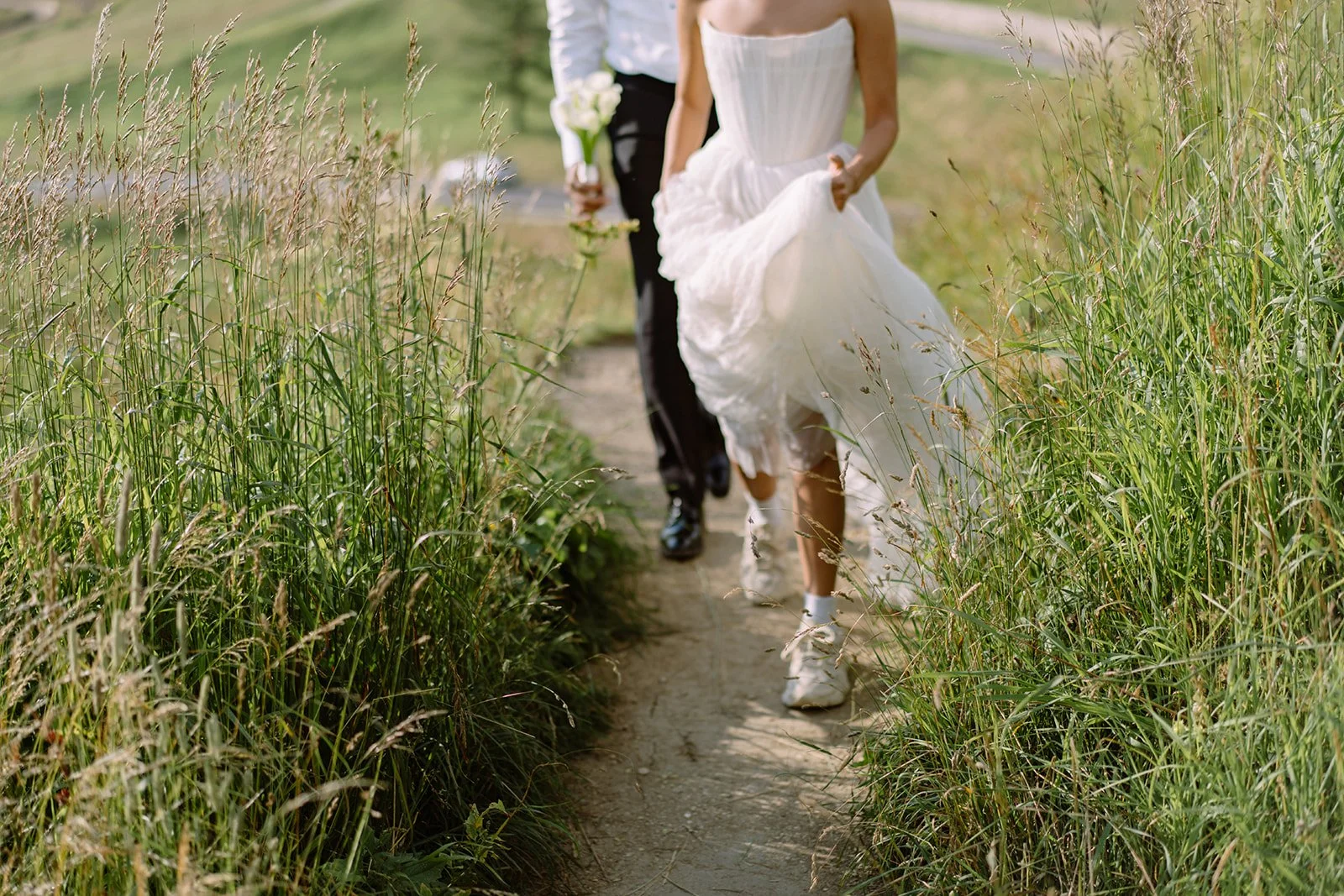 A bride and groom walking along a narrow dirt path through tall grass on their wedding day.