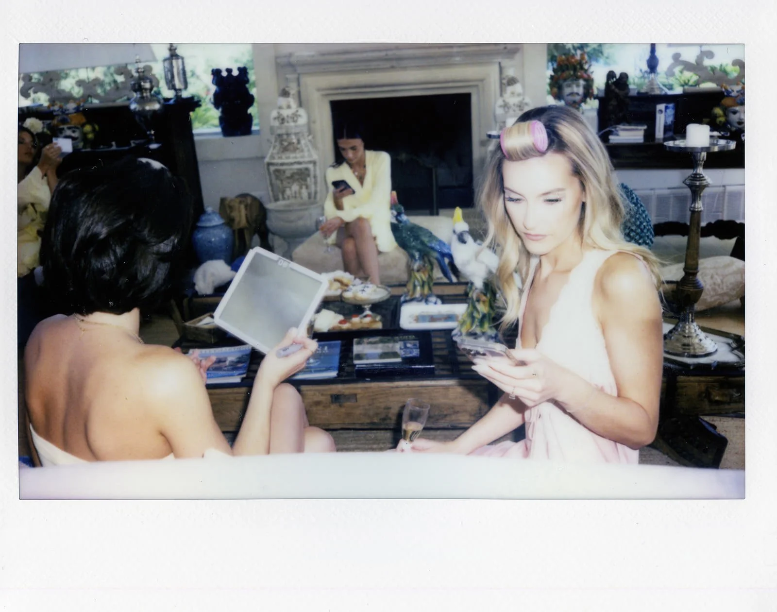 Group of women gathered in a living room with a fireplace, some looking at their phones, while one is looking at a photo album. The table in front has decorative parrots and a champagne glass, with more decorations and furniture in the background.