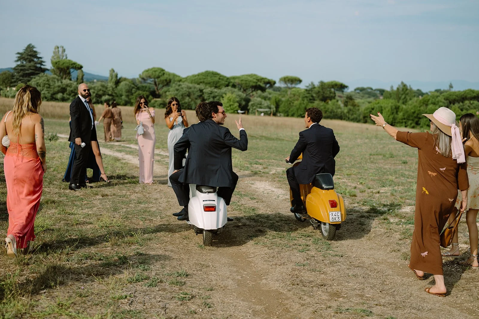 People gathered outdoors in a grassy field, some on scooters and others standing, with trees and hills in the background, during daytime.
