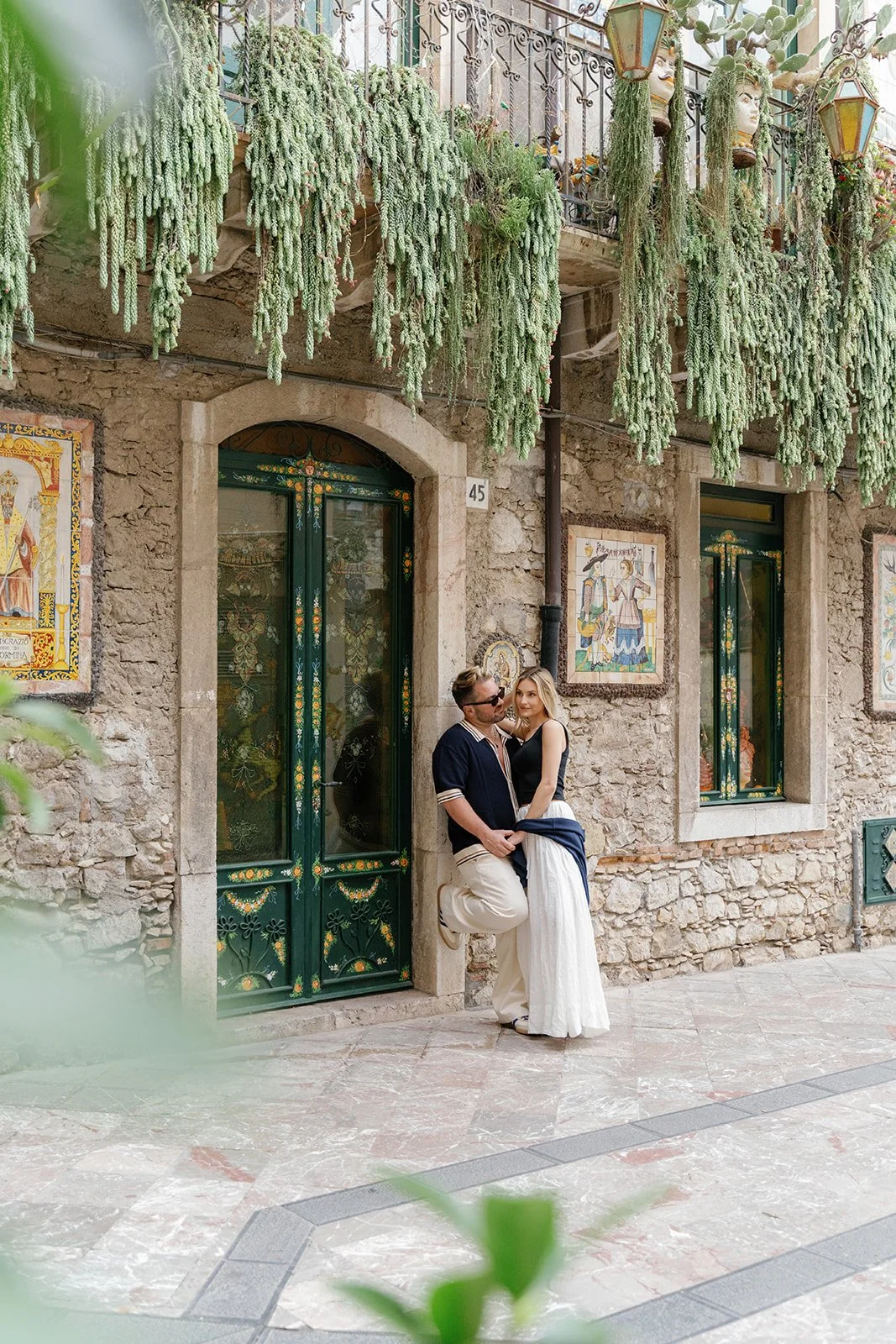 A couple standing close to each other outside a stone building with green doors decorated with floral patterns, surrounded by hanging plants and colorful tiles on the wall.
