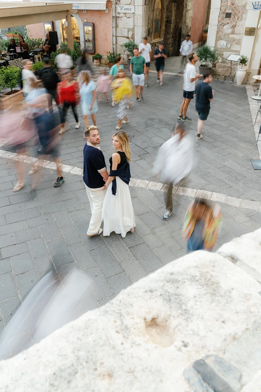 A couple standing close together, holding hands, in the middle of a busy cobblestone street with blurred pedestrians around them. The woman is wearing a white flowing skirt and a black top, while the man is dressed in a navy blue sweater and white pa