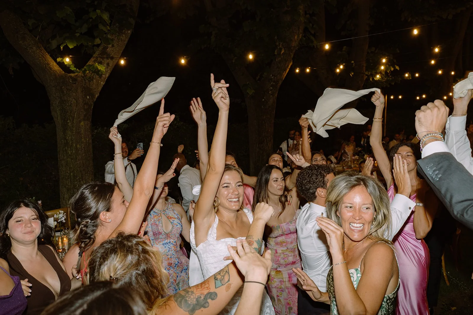 People dancing and celebrating at an outdoor night party under string lights, smiling and raising their hands.