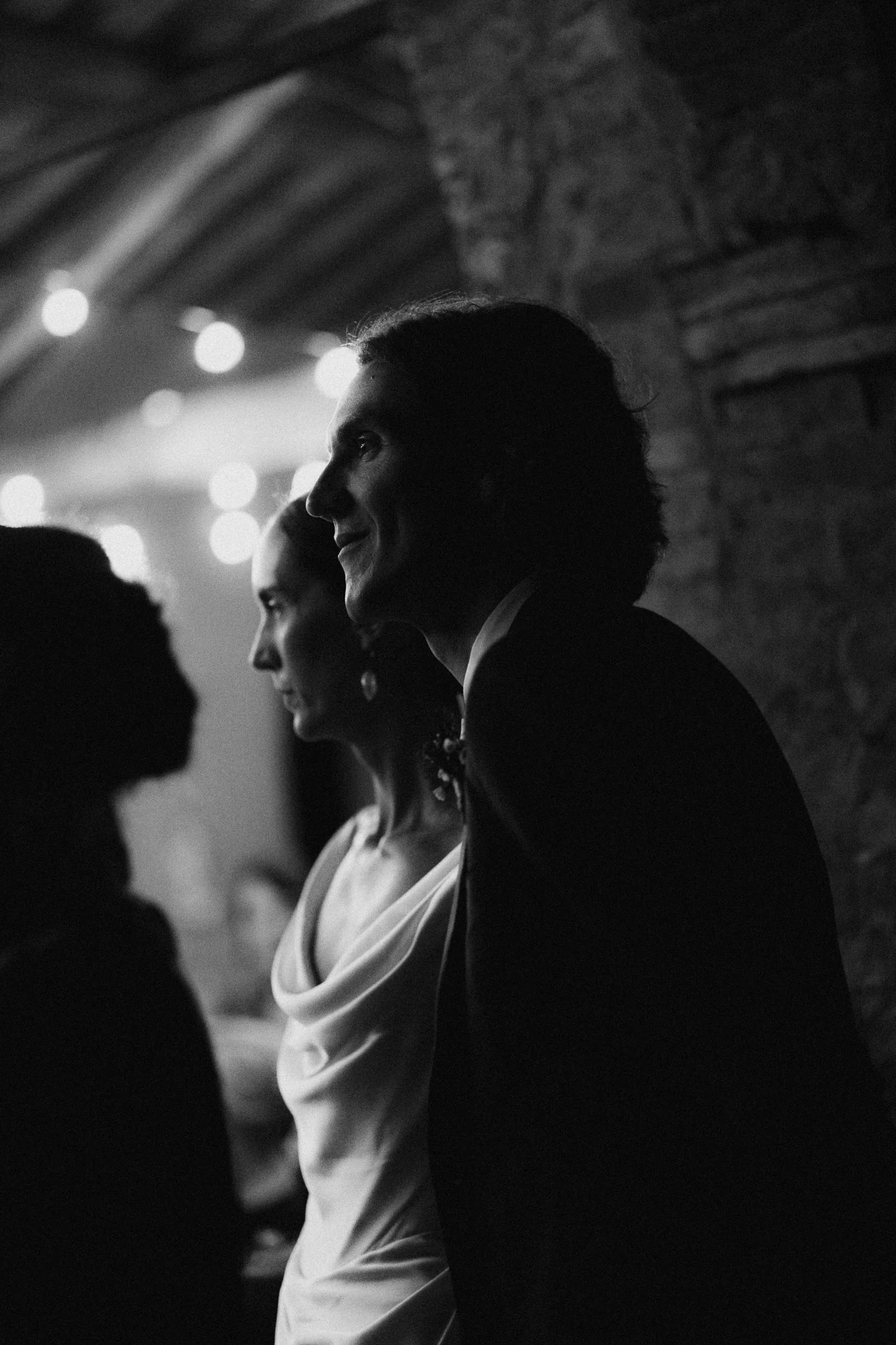 Black and white photo of three people in formal attire, standing in a dimly lit room with string lights, profile view, smiling and engaging in conversation.