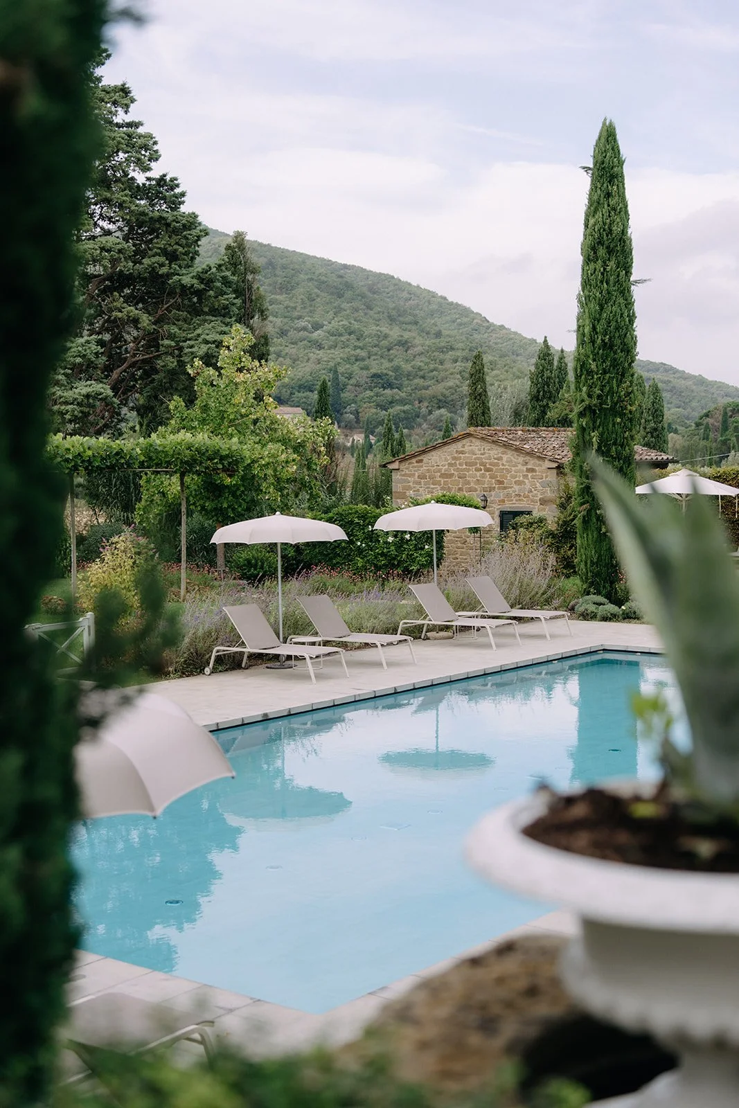 Swimming pool with white lounge chairs and umbrellas surrounded by lush greenery and tall trees, with a rustic stone building and hills in the background.