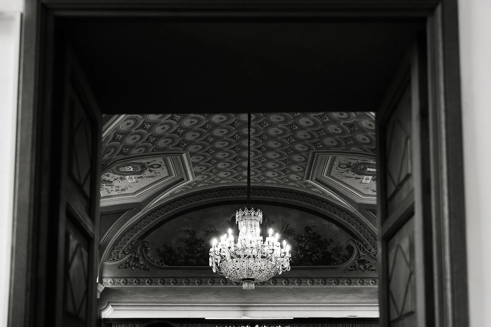 View through open double doors showing ornate ceiling with floral and geometric patterns and a large chandelier in a grand room.