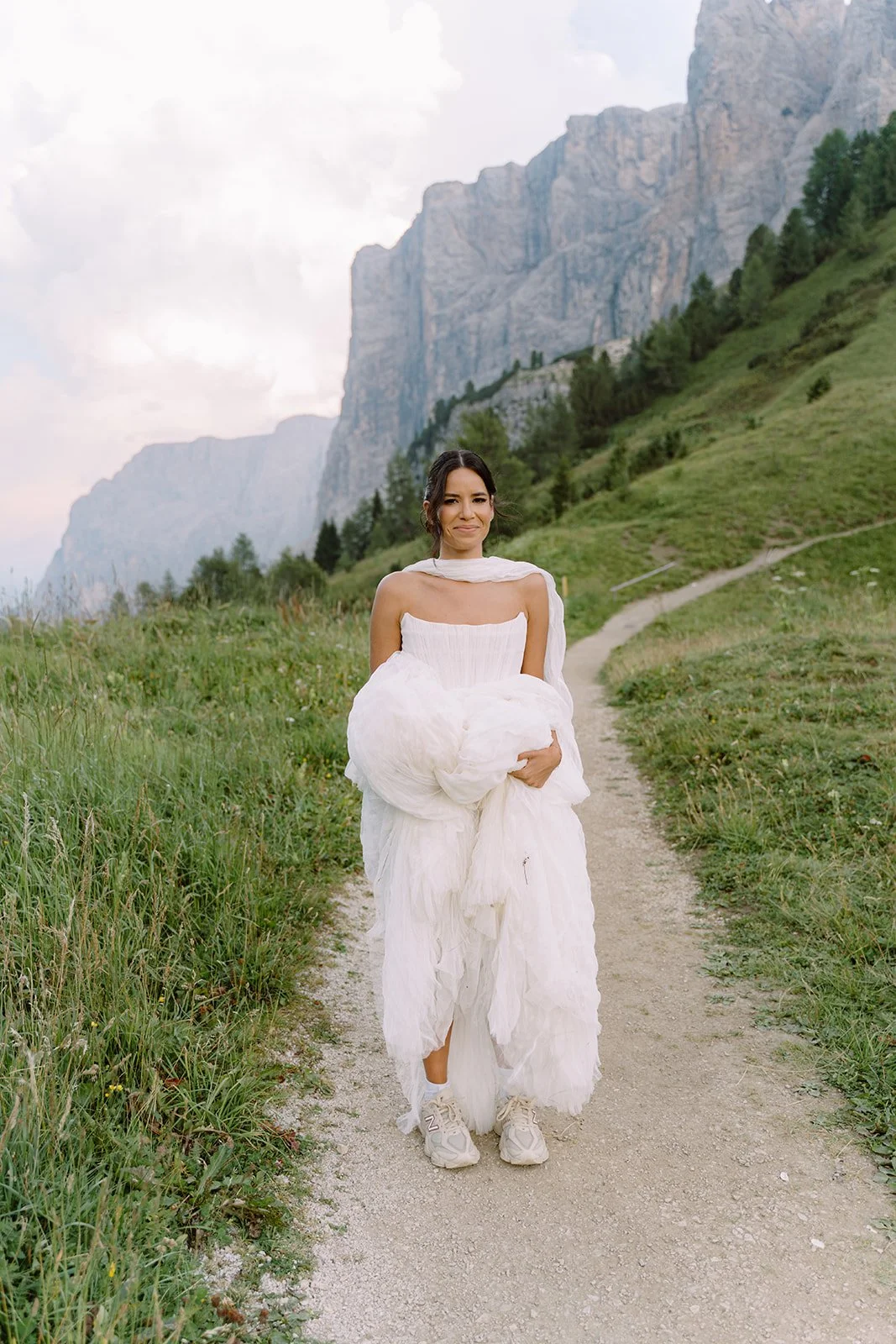 Woman in a white dress and sneakers walking on a trail in a lush green mountain landscape.