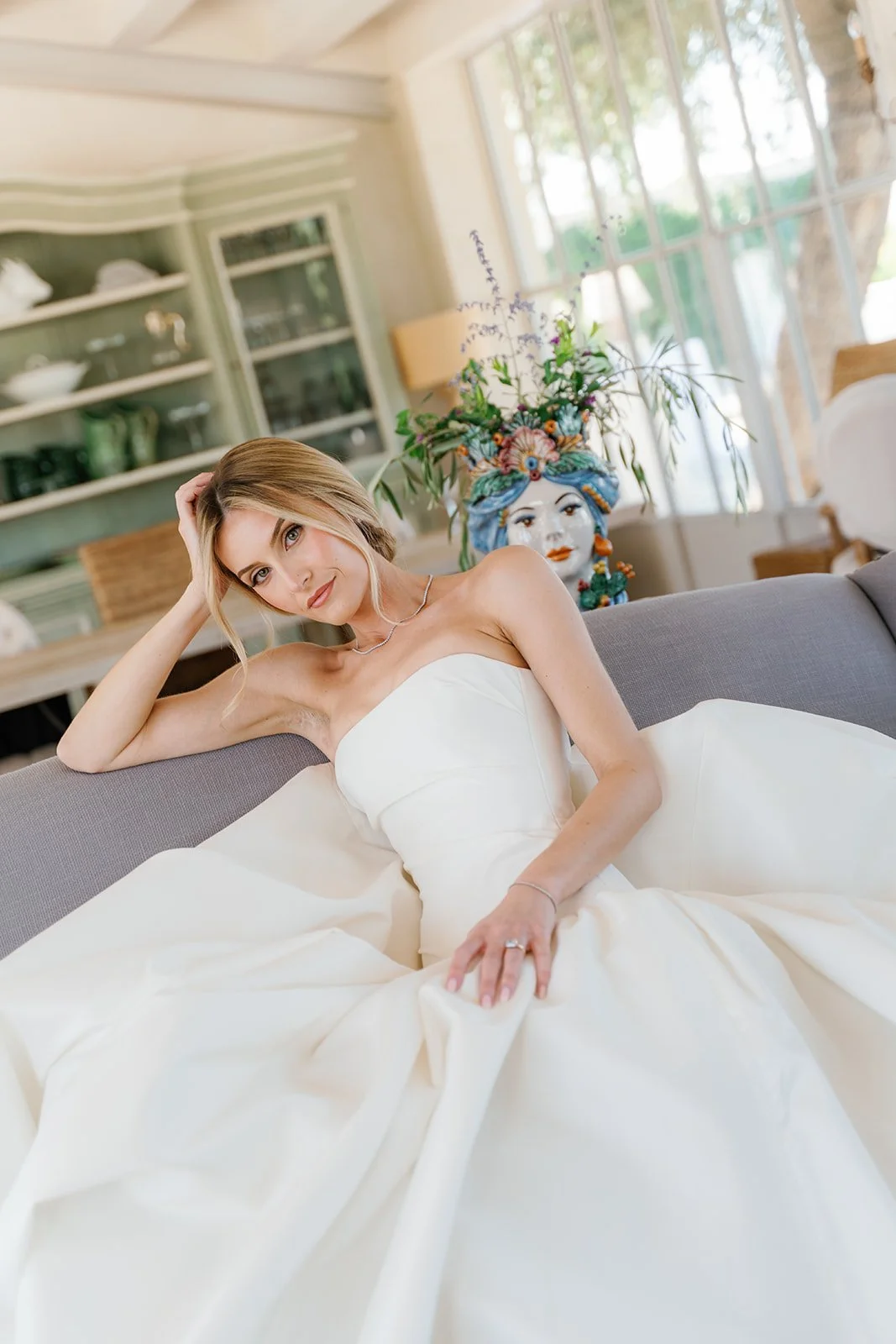 A woman in a white strapless wedding dress lying on a gray sofa in a bright room with large windows.