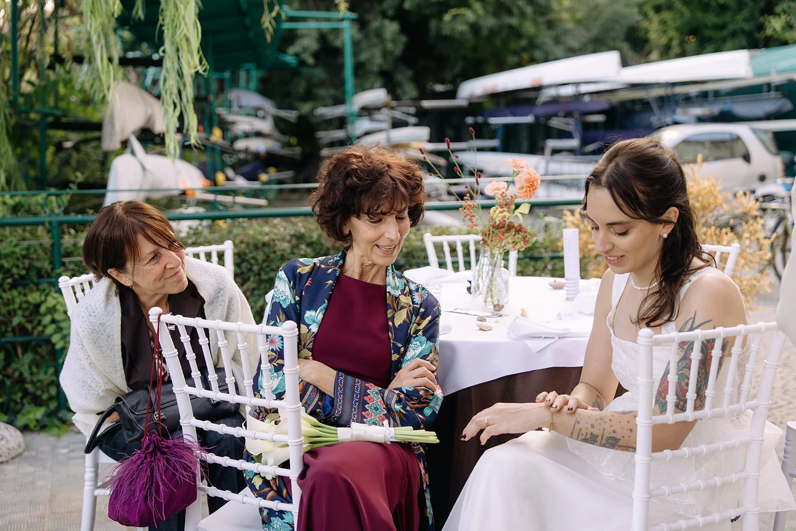A wedding scene with three women sitting at a table outdoors. The bride has dark hair, a white dress, and tattoos, and is showing her hand to a woman with curly hair in a floral dress. An elderly woman with short brown hair and a woman with short red