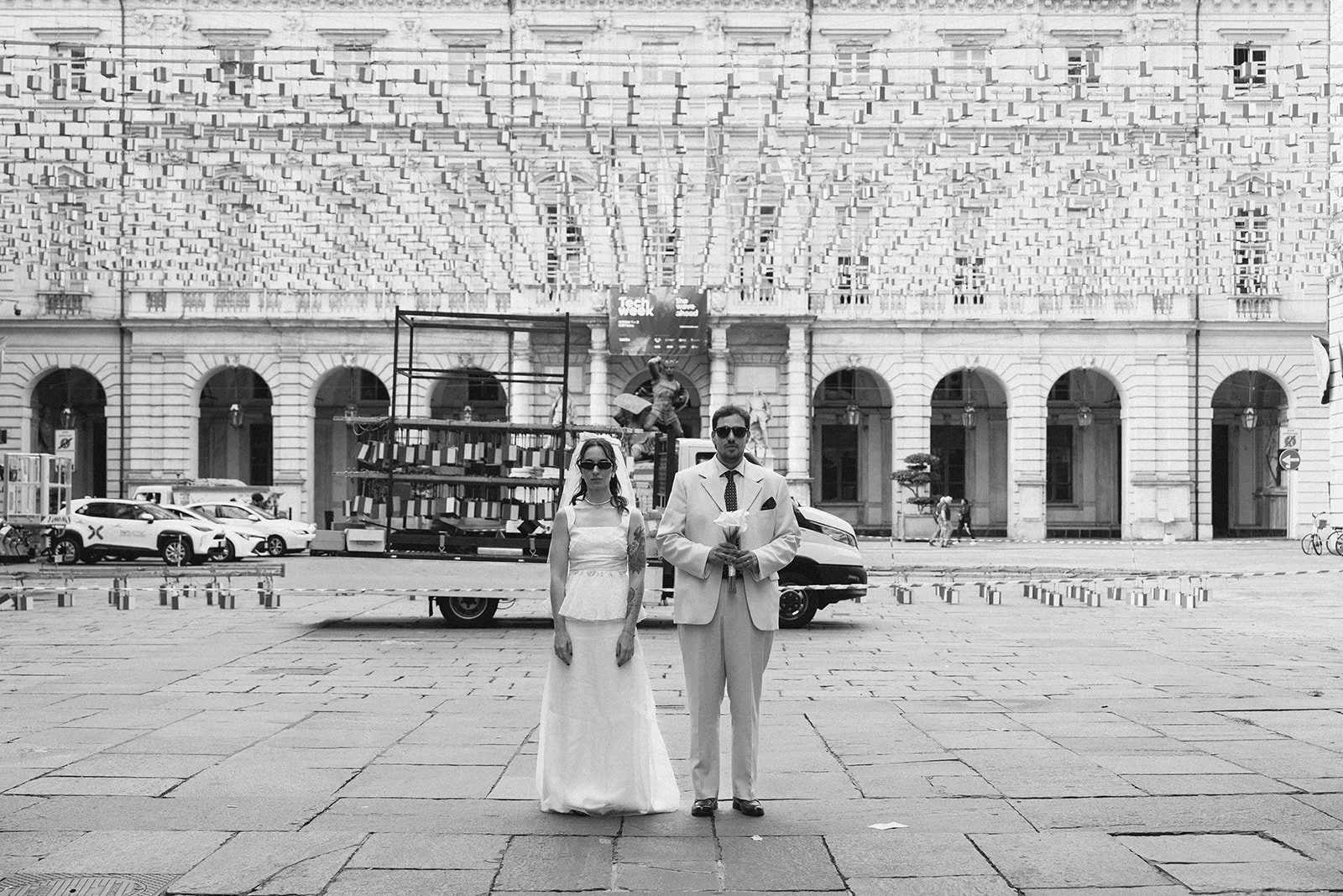 A black and white photo of a man and woman in wedding attire standing on a city street with buildings, parked cars, and a truck in the background.