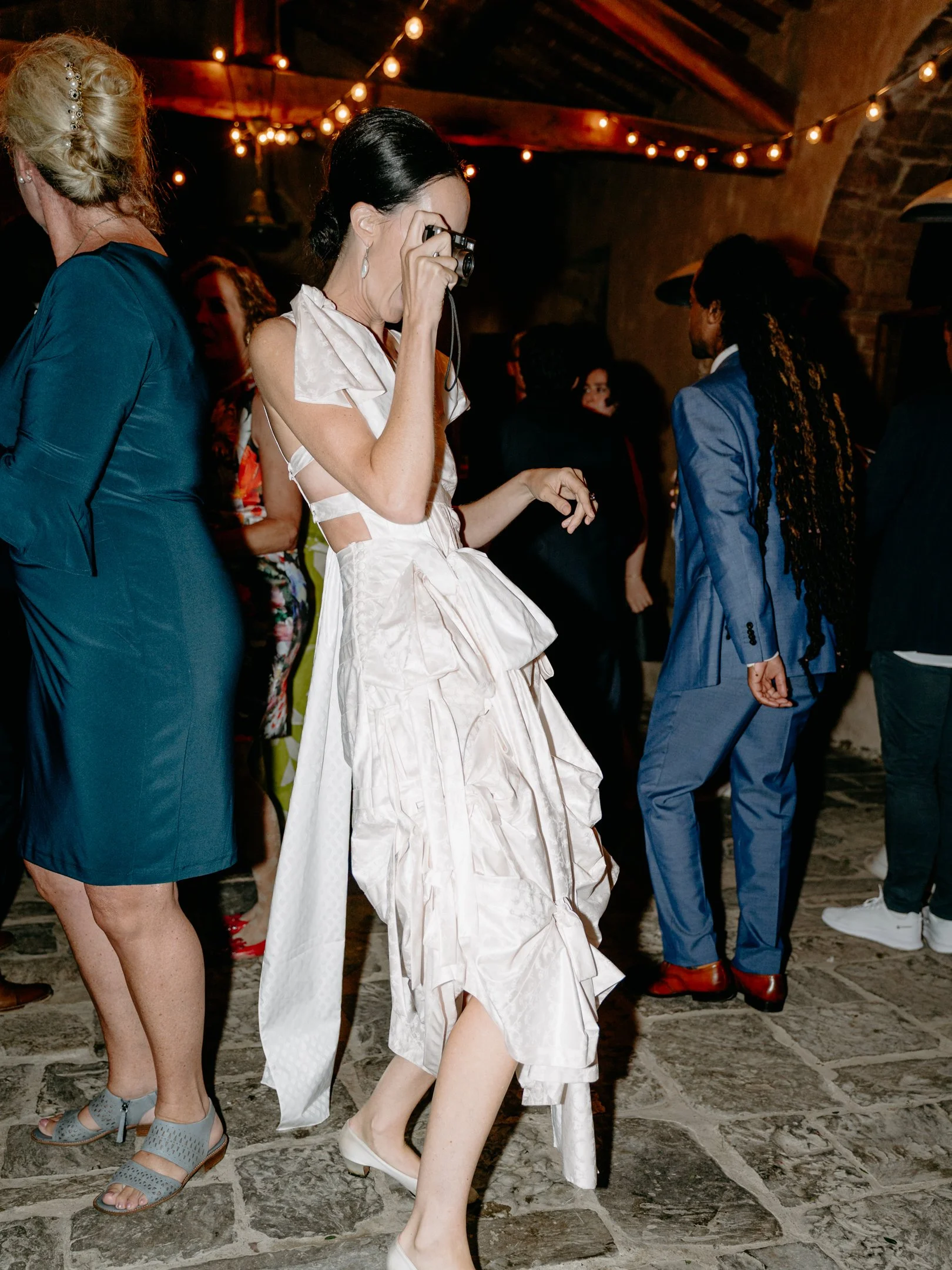 A woman in a cream-colored, ruffled dress is taking a photo at a social event under string lights in a rustic setting.