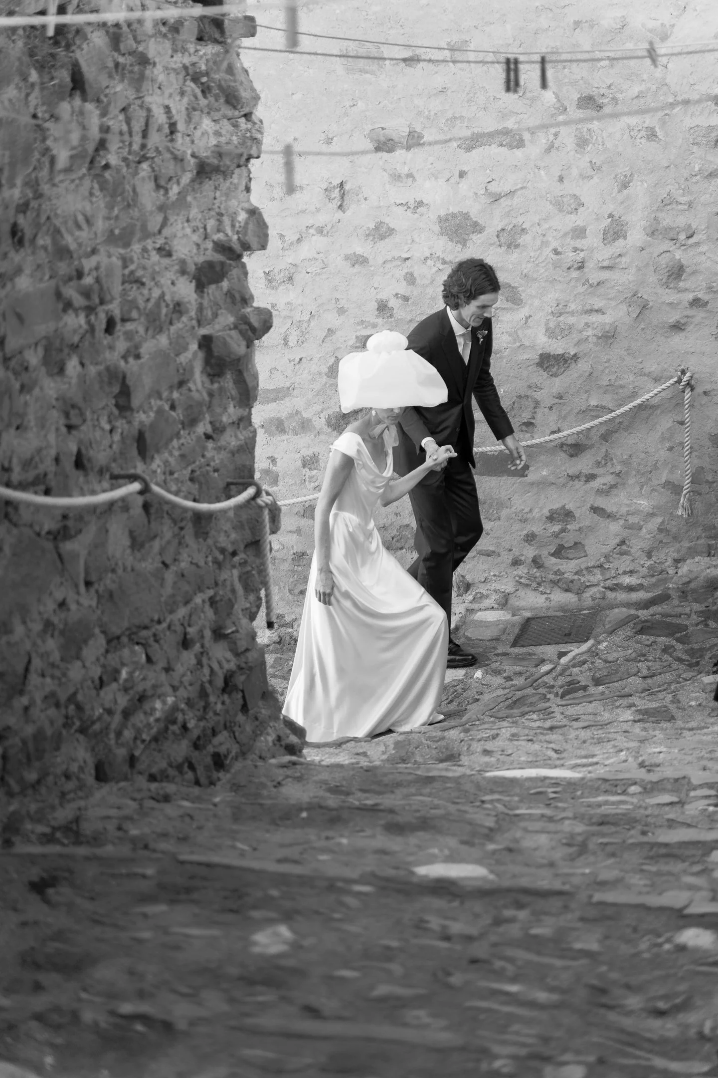 A black-and-white photo of a bride and groom walking up a cobblestone alleyway. The bride wears a white dress and a large hat, and the groom is in a dark suit. They are holding hands as they ascend the stairs.