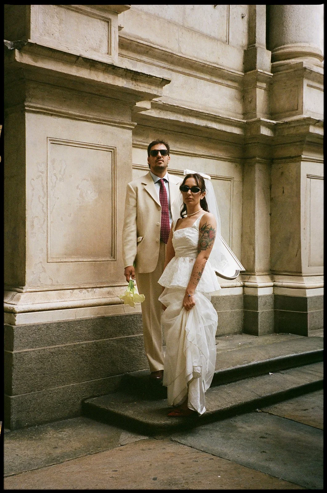 A couple dressed in wedding attire standing outside on a stone sidewalk in front of a historic stone building. The man is wearing a beige suit with a white shirt, red tie, and sunglasses, holding a bouquet of flowers. The woman is wearing a white wed