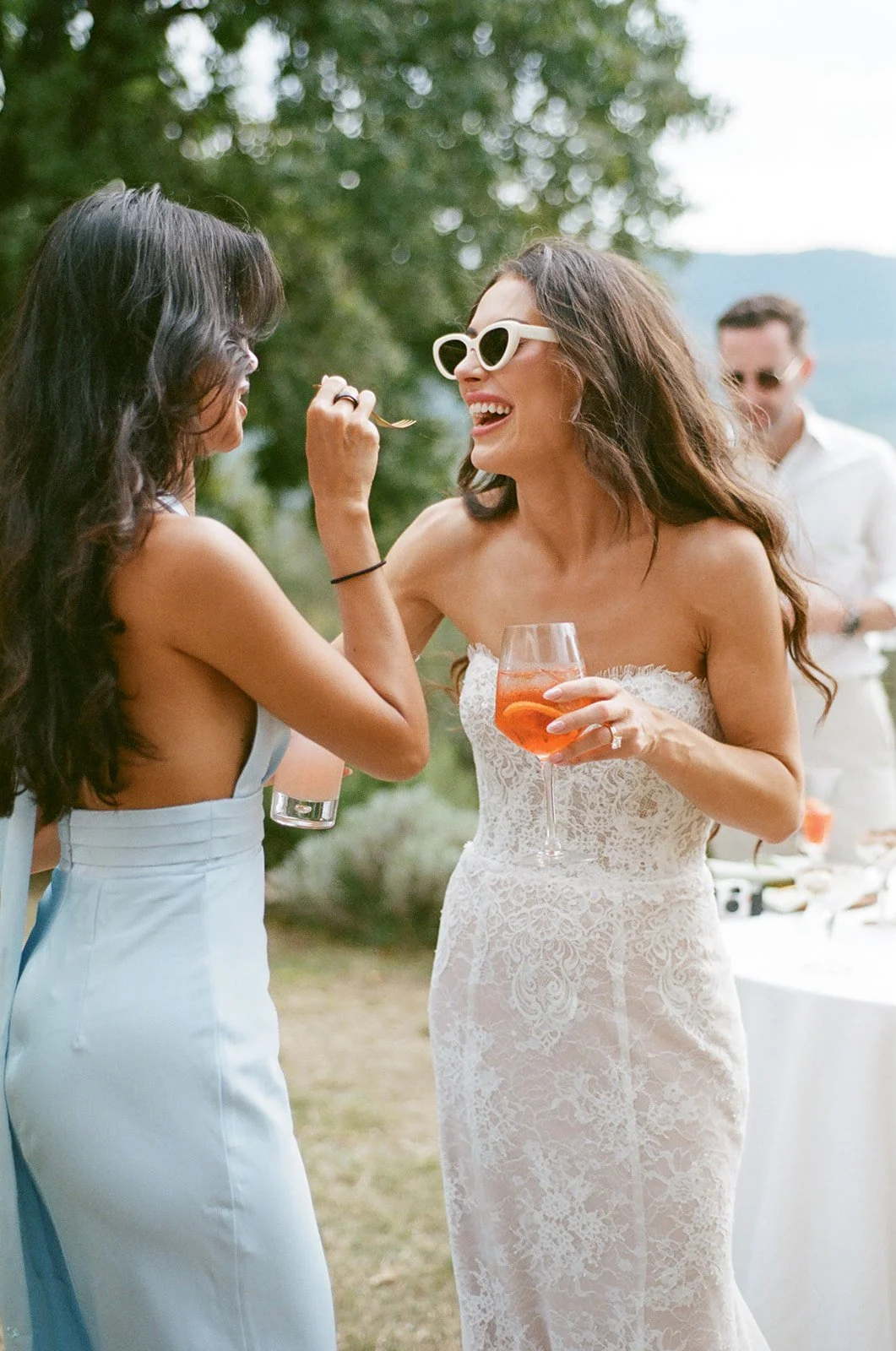 Two women at an outdoor wedding, one feeding the other with a fork, both smiling and enjoying the moment. One woman is wearing a white dress with lace details and sunglasses, holding a glass of pink cocktail. The other woman is in a strapless pastel 