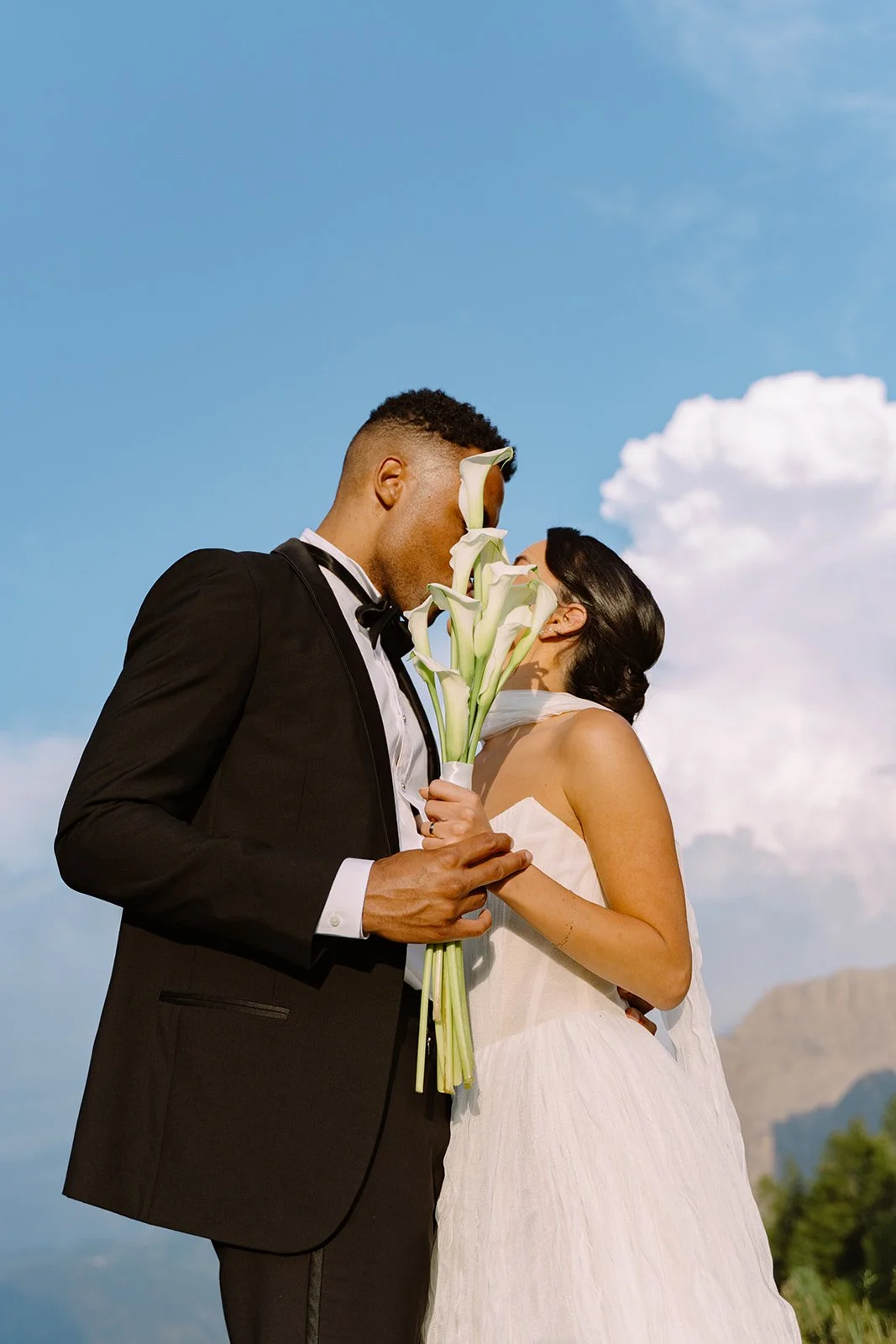 A couple dressed in wedding attire, holding a bouquet of calla lilies, sharing a kiss outdoors against a blue sky with clouds and mountains in the background.