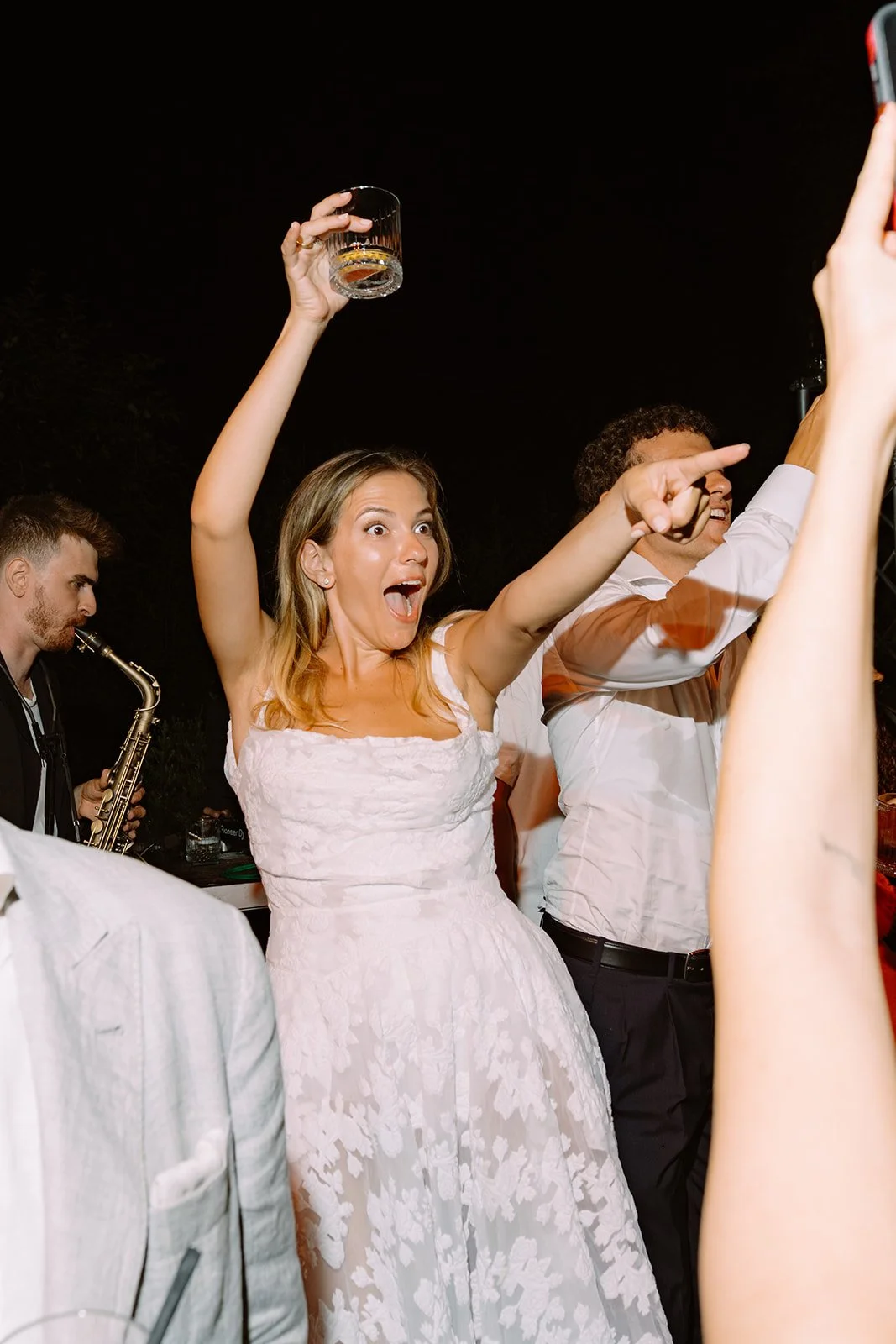 Woman in white dress celebrating with a glass of alcohol at a party, with people around her and a musician playing saxophone in the background.