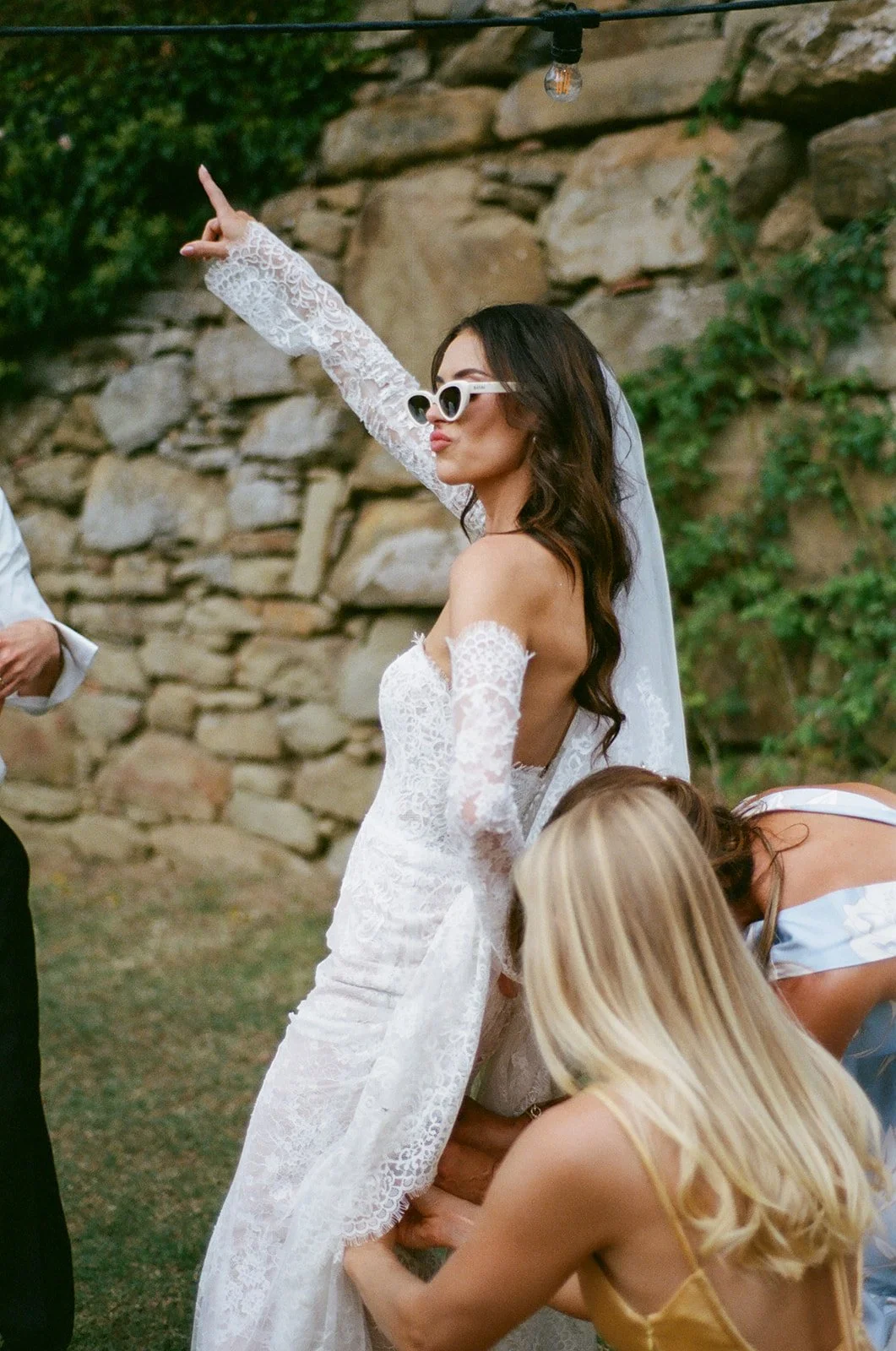 A woman in a white lace wedding dress and sunglasses strikes a confident pose outdoors at a wedding.