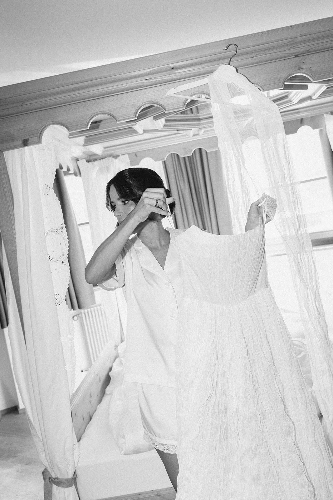 A woman in white pajamas holding a dress while standing in front of a mirror in a bedroom with curtains and a four-poster bed.