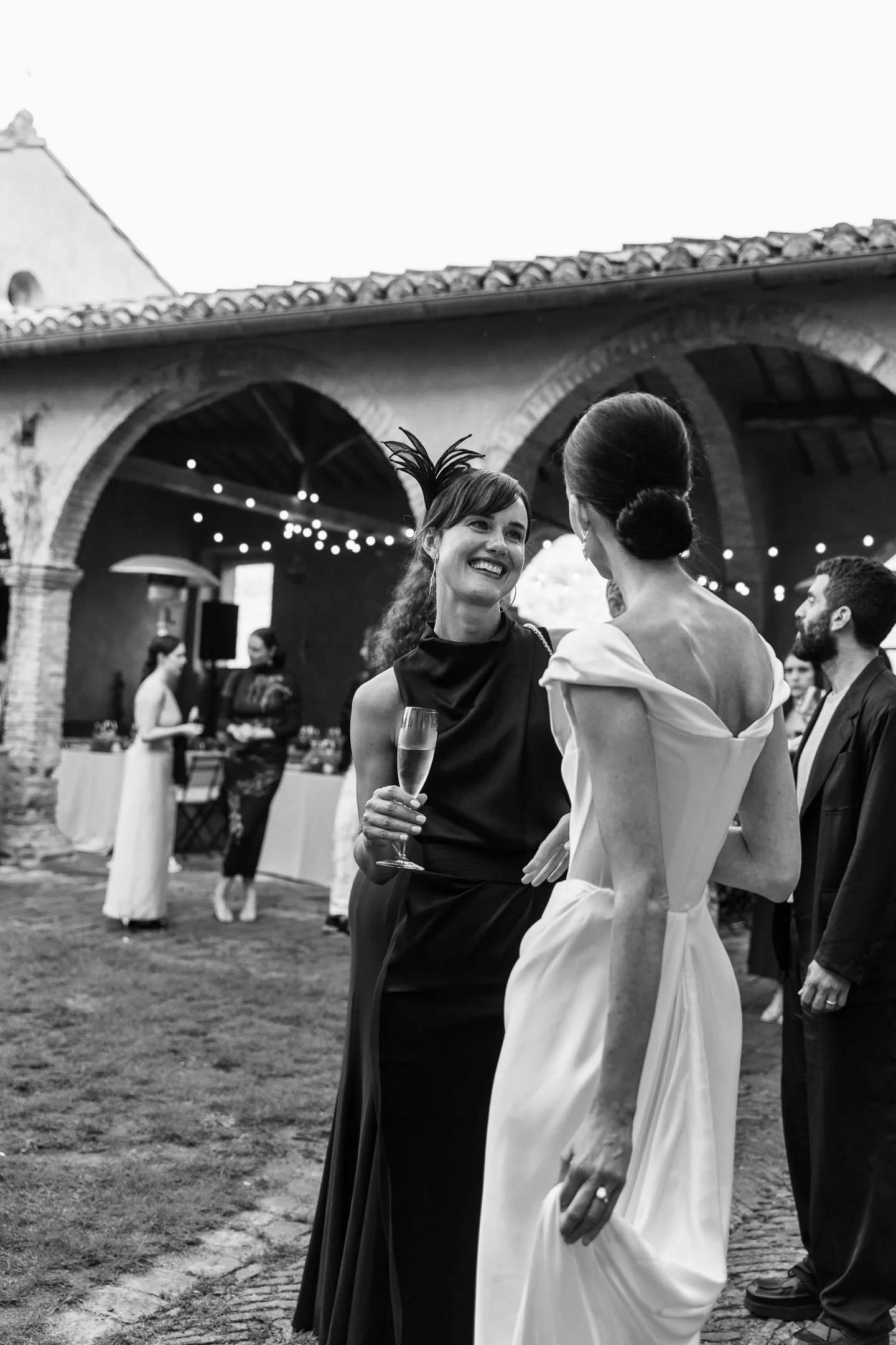 Black and white photo of women at a wedding reception, chatting, with one holding a champagne glass. The woman facing the camera wears a dark dress with a fascinator, while the bride wears an off-the-shoulder gown. Background features other guests an