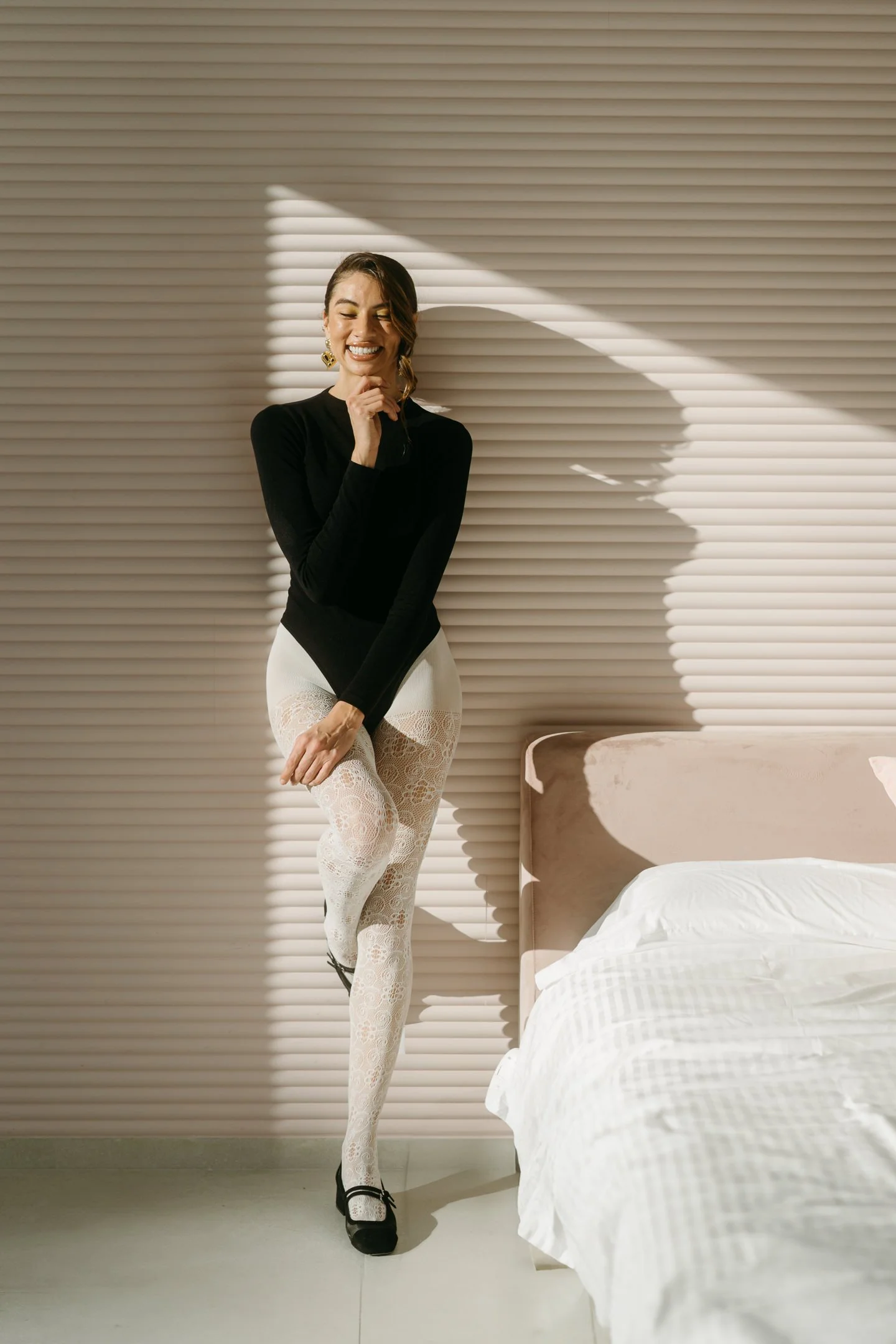 A woman in black top and white lace tights standing in a room with beige horizontal slat wall, smiling, with her eyes closed, standing near a bed with white sheets.