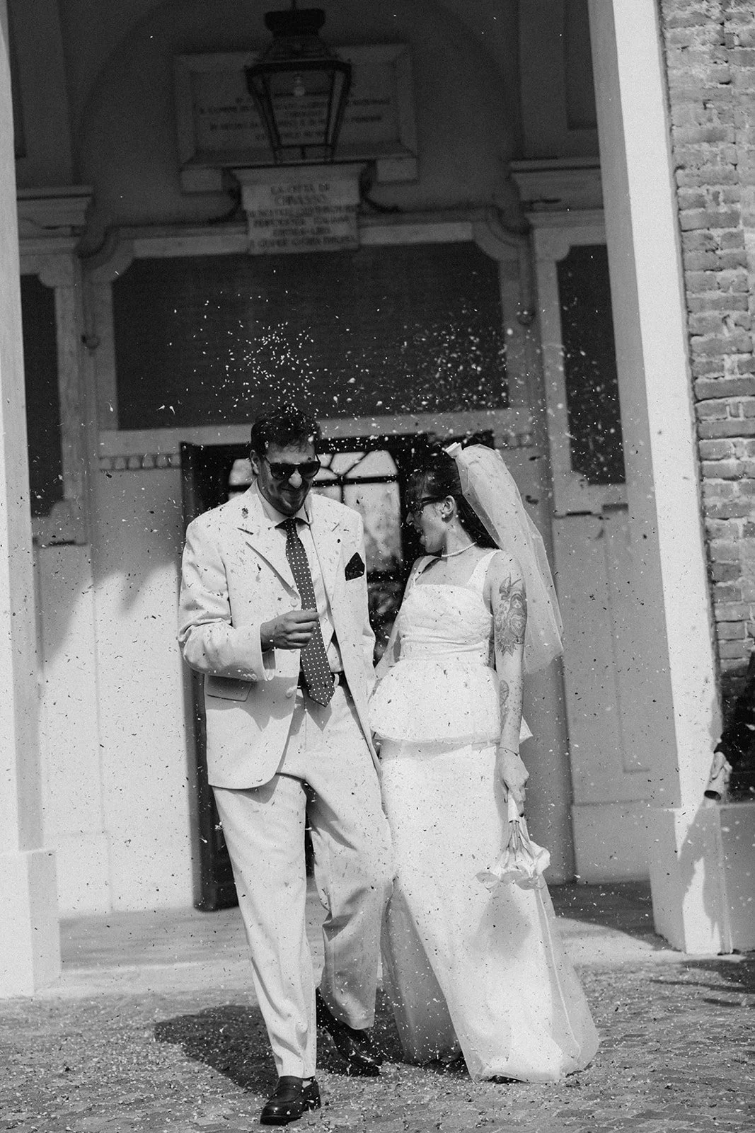 A couple in wedding attire celebrating outside a building, throwing confetti into the air. The groom is wearing sunglasses, a light-colored suit, and a polka-dot tie. The bride is wearing a strapless wedding gown with tattoos on her arm, and she hold