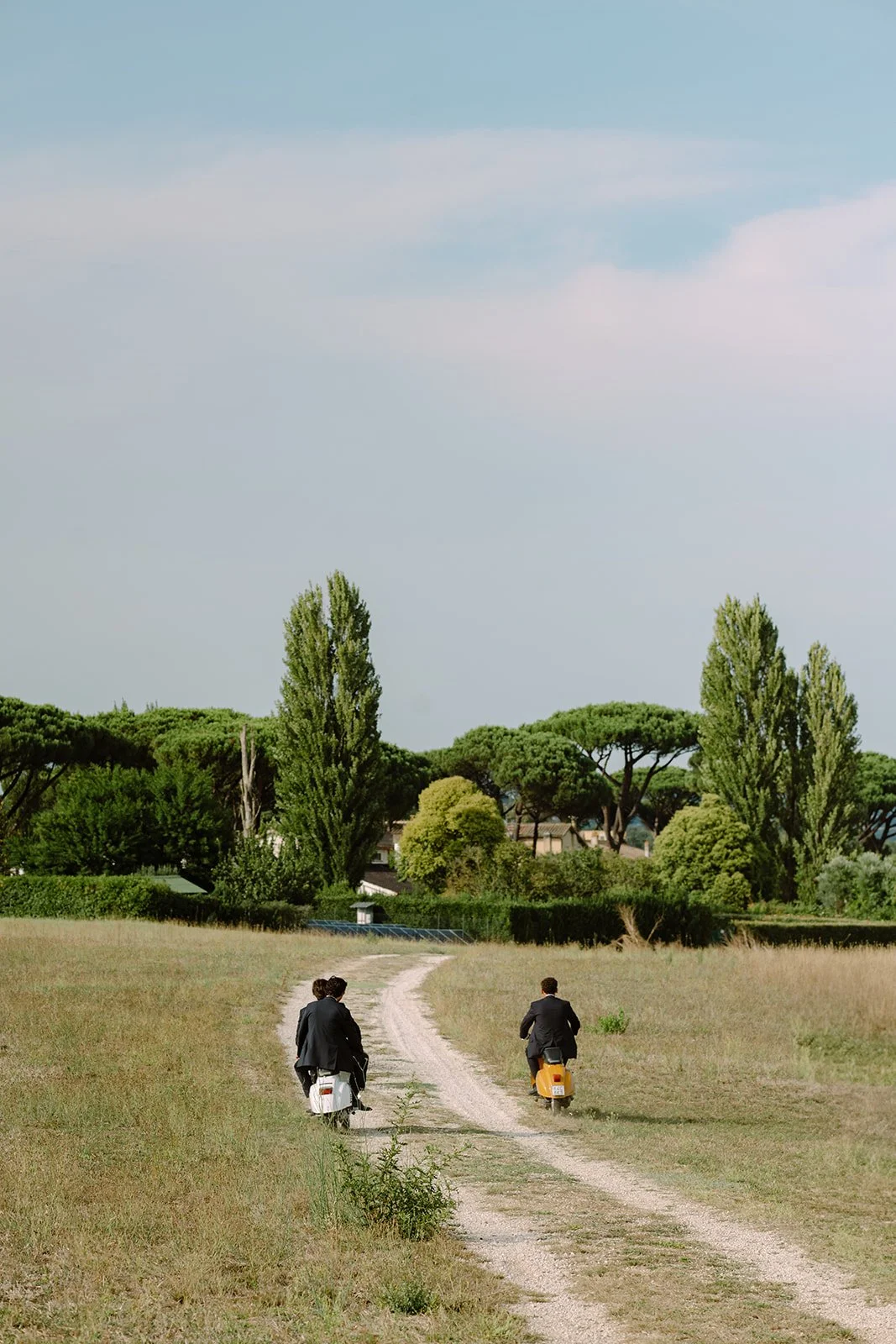 Two men in suits riding scooters on a dirt path through a grassy field with trees in the background.
