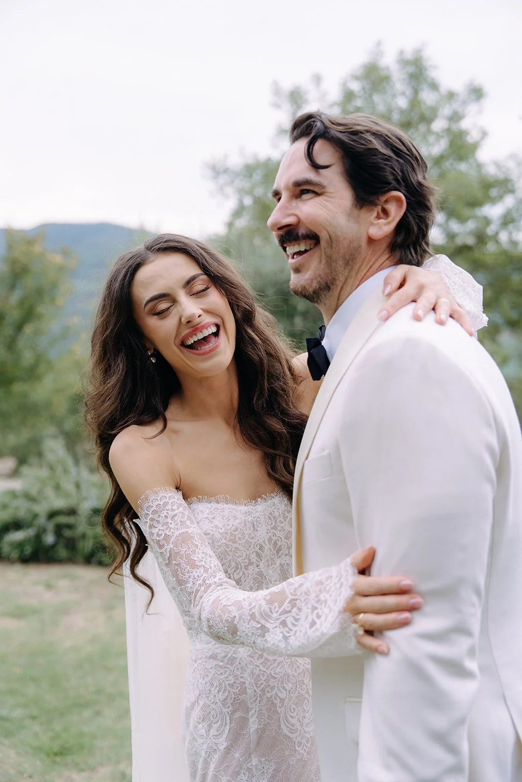 A bride and groom sharing a joyful moment outdoors, with the bride laughing and the groom smiling, surrounded by greenery.