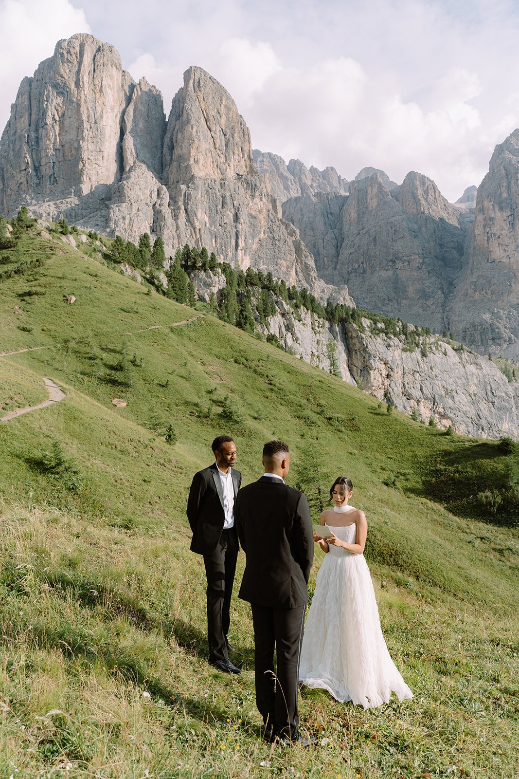 A wedding ceremony taking place outdoors on a grassy hillside with mountains in the background. The bride in a white dress is reading vows while two groomsmen in black suits stand nearby.