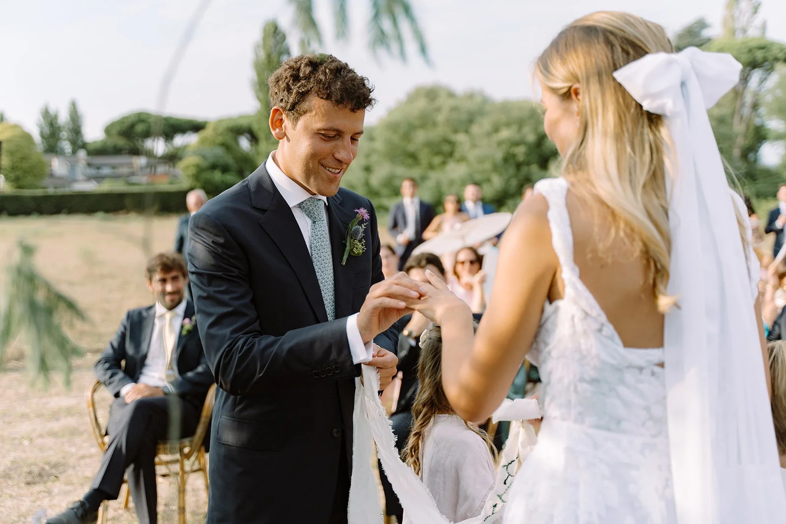 A couple exchanging rings during an outdoor wedding ceremony with guests in the background.
