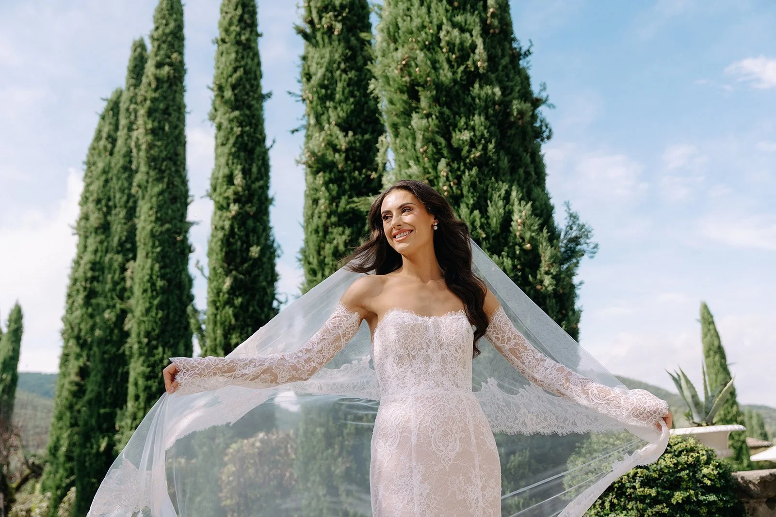 A bride in a white lace wedding dress with off-the-shoulder sleeves standing outdoors, smiling, with tall green cypress trees and a clear blue sky in the background.