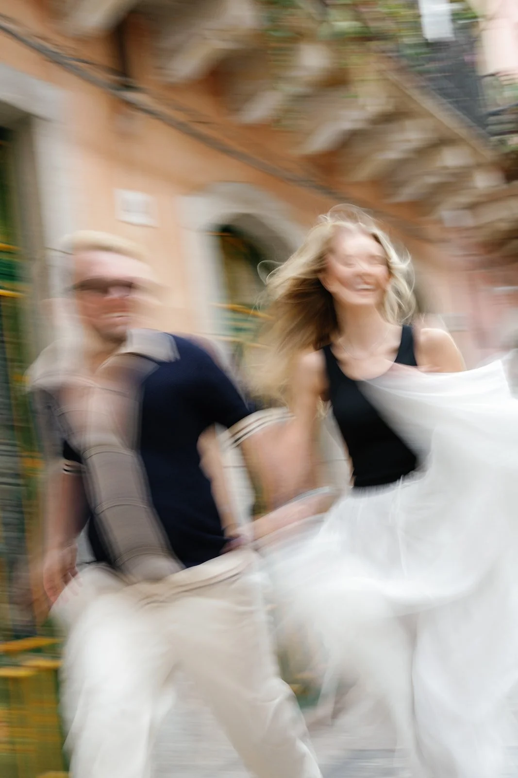 A blurred photo of a smiling woman with blonde hair and a man in the background, walking outdoors near buildings.