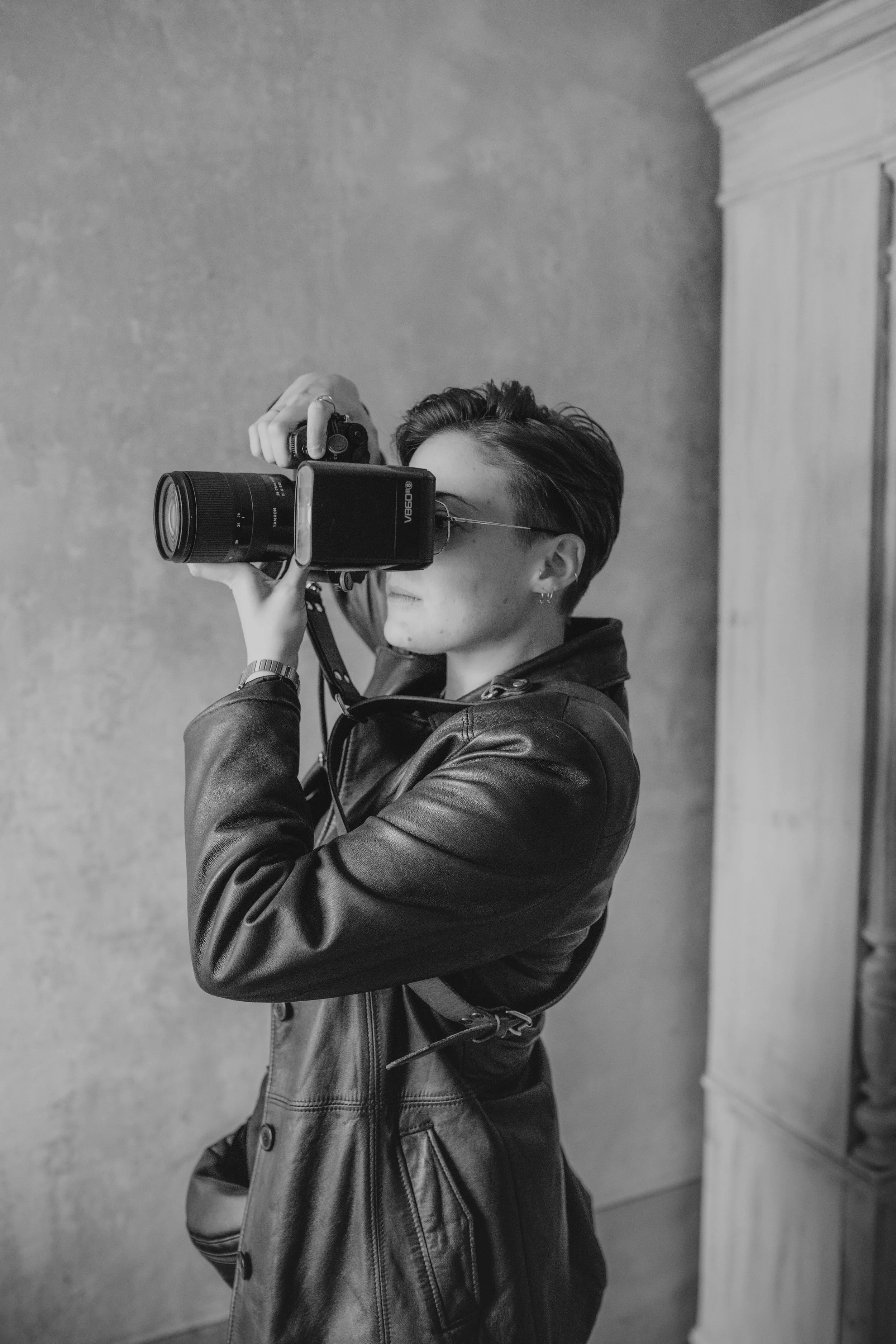 A person with short hair, wearing sunglasses and a leather jacket, is taking a photo with a camera.