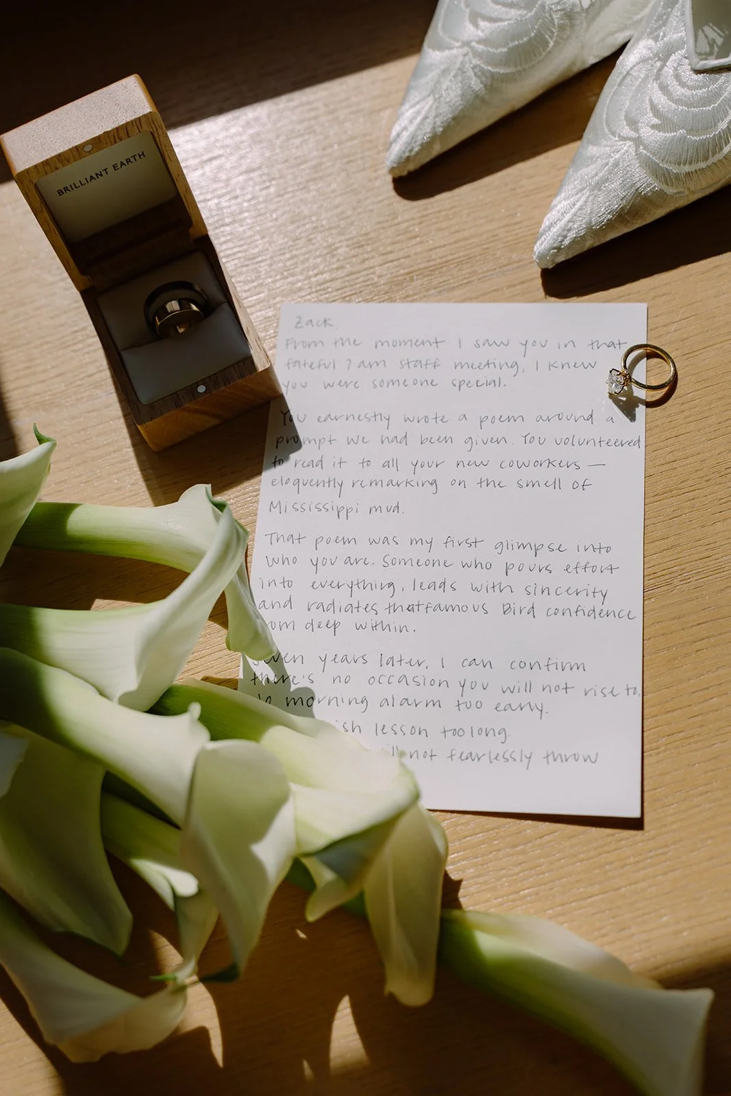 A close-up of a wooden table with a white lily flower, a handwritten note, a ring in an open box labeled 'Brilliant Earth,' and a pair of white embroidered napkins.