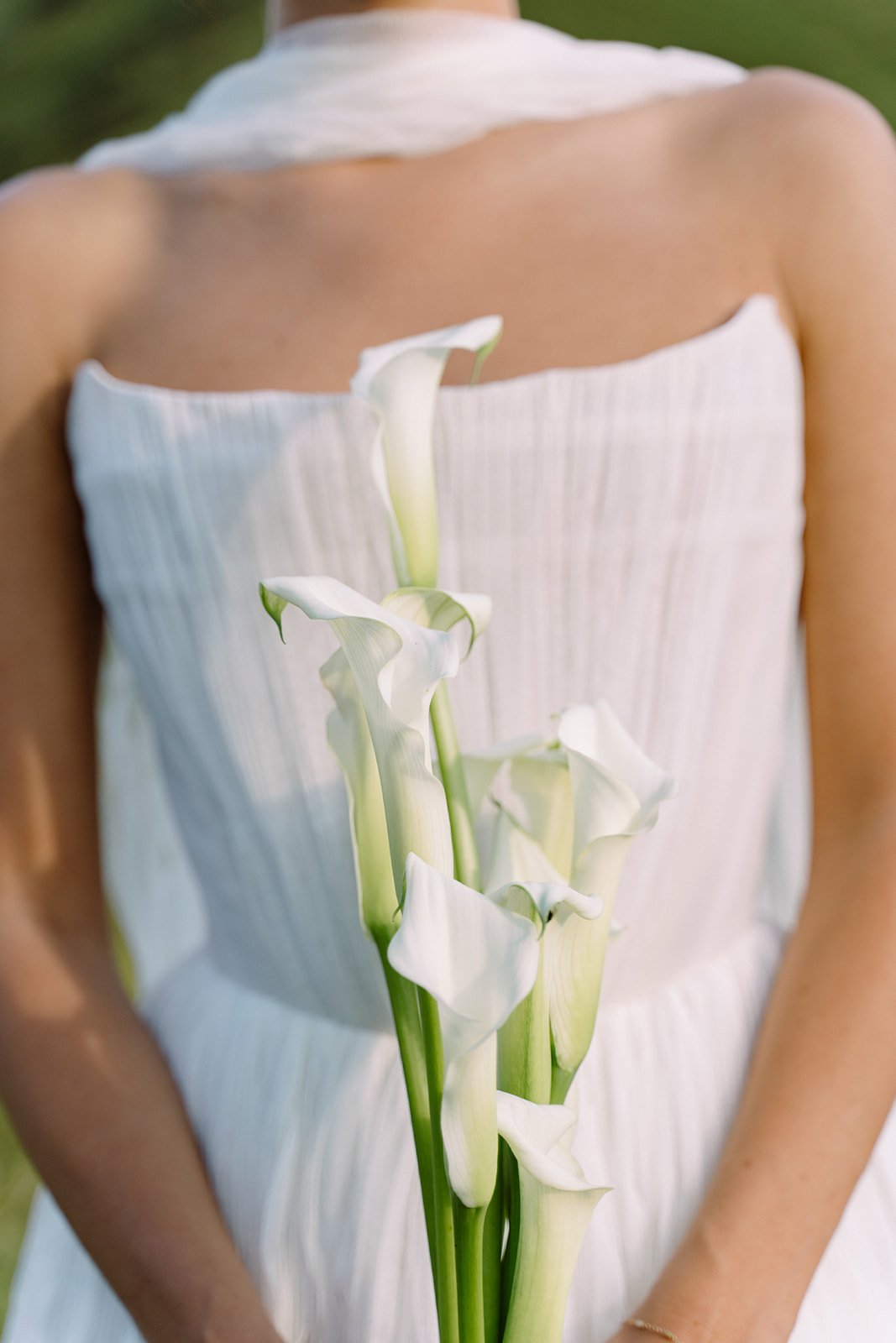 Close-up of a woman in a white dress holding white calla lilies against a blurred green outdoor background.