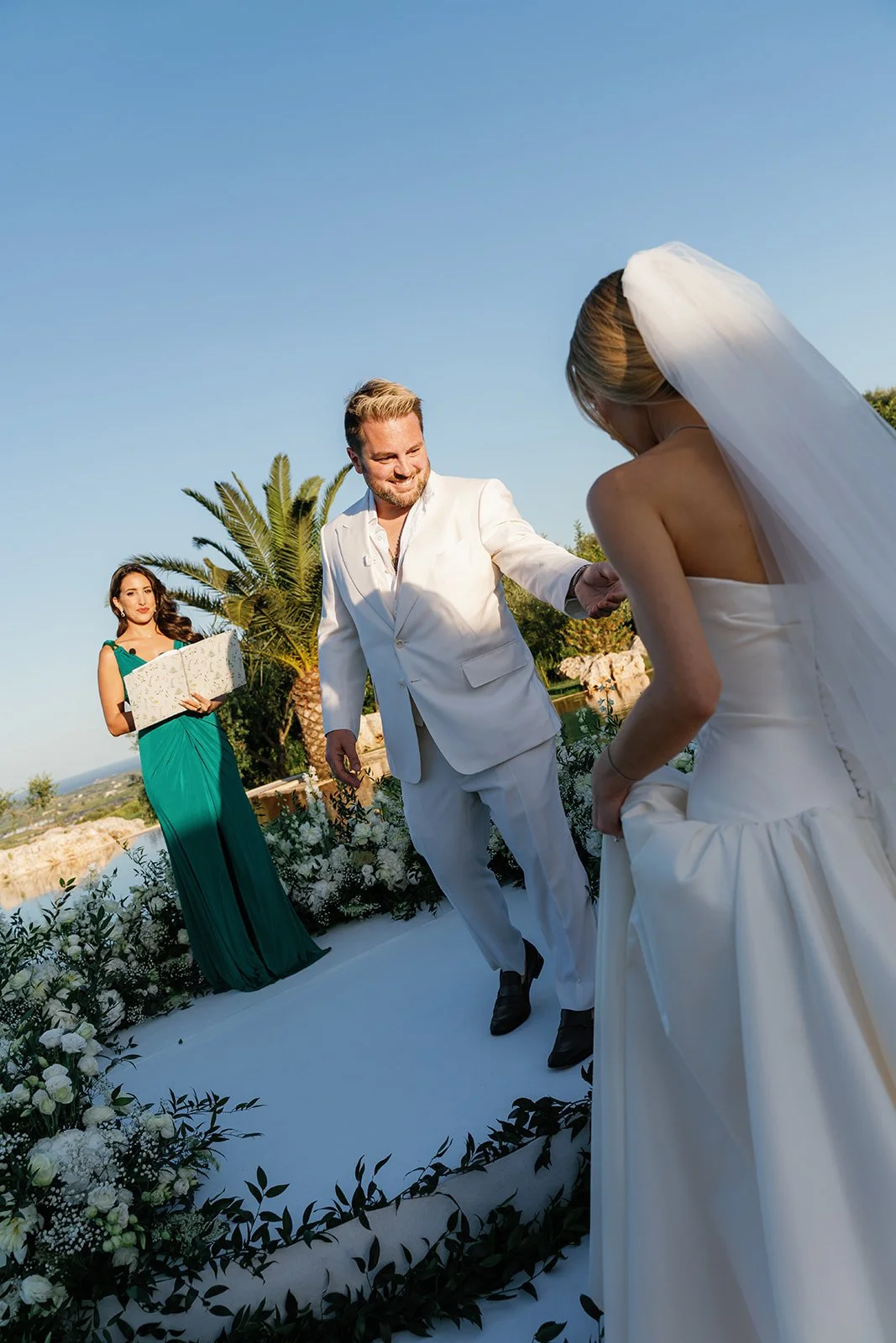A wedding ceremony outdoors with a bride, groom, and officiant, surrounded by flowers and greenery on a sunny day.