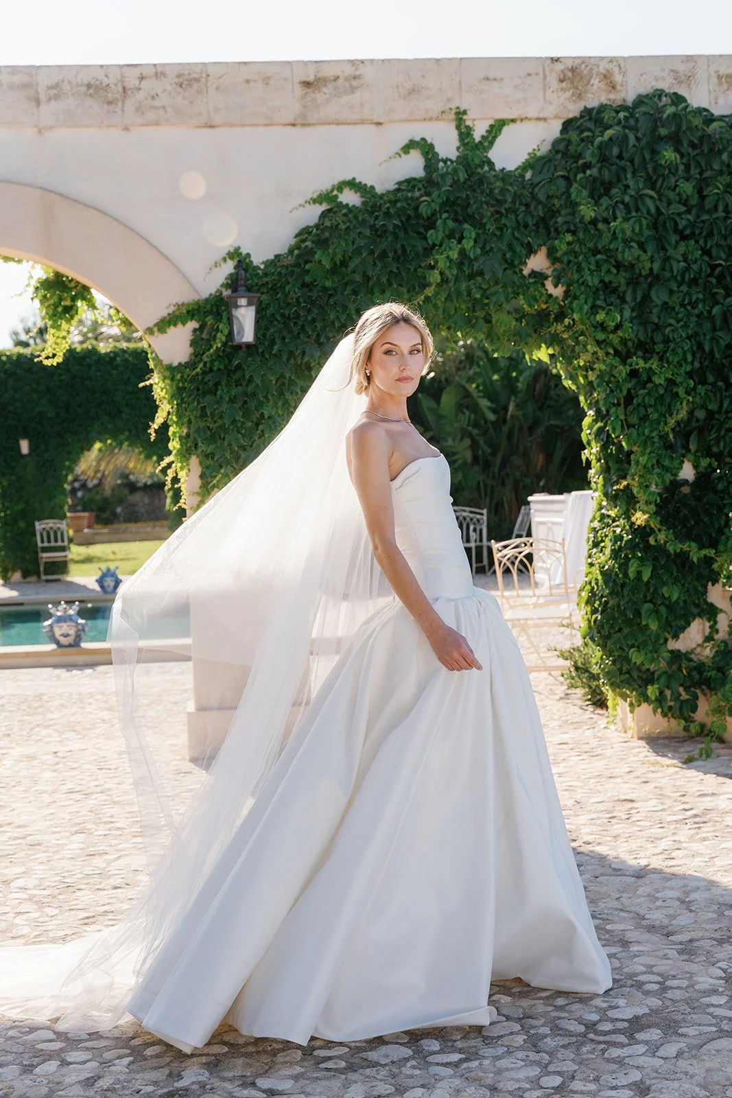 A woman in a white wedding gown standing outdoors under an arched stone structure covered with green ivy. She is wearing a veil and has a confident expression.