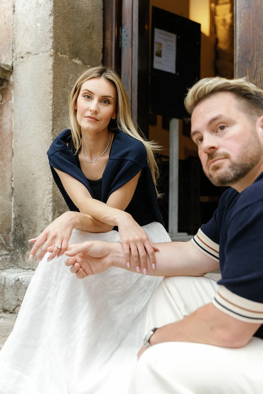 A woman with long blonde hair and a man with light brown hair and a beard sitting outside a building with stone walls. The woman is looking directly at the camera, while the man looks slightly away. They are holding hands, and the woman is wearing a 