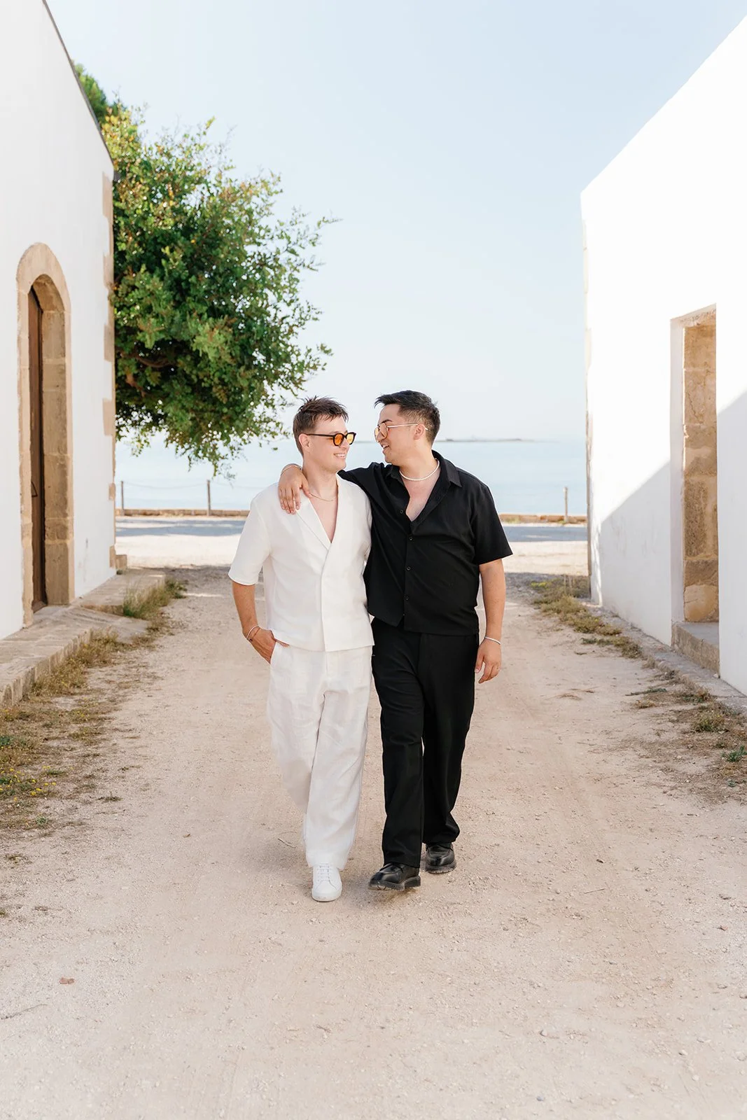 Two people walking together, one with short hair in a white outfit and the other with glasses in a black outfit, smiling and embracing on a dirt pathway during daytime, with a tree on the left and the ocean in the background.