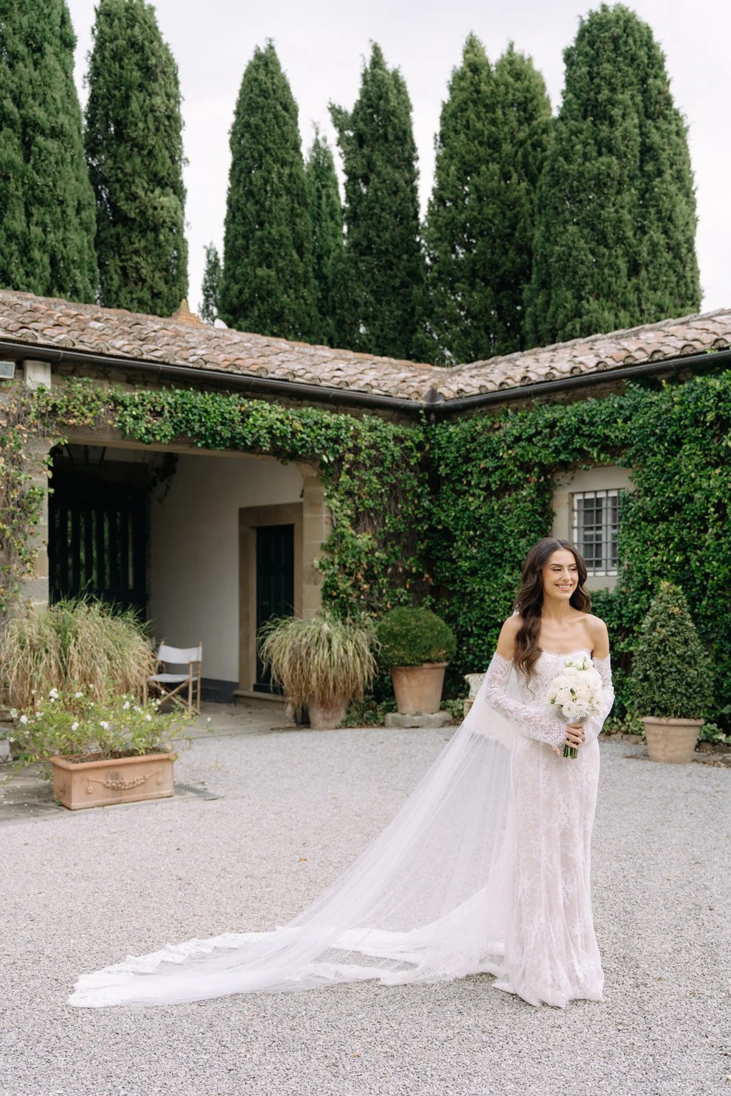 Bride in a white lace wedding gown holding a bouquet of white flowers standing outdoors in a courtyard with potted plants and lush greenery.