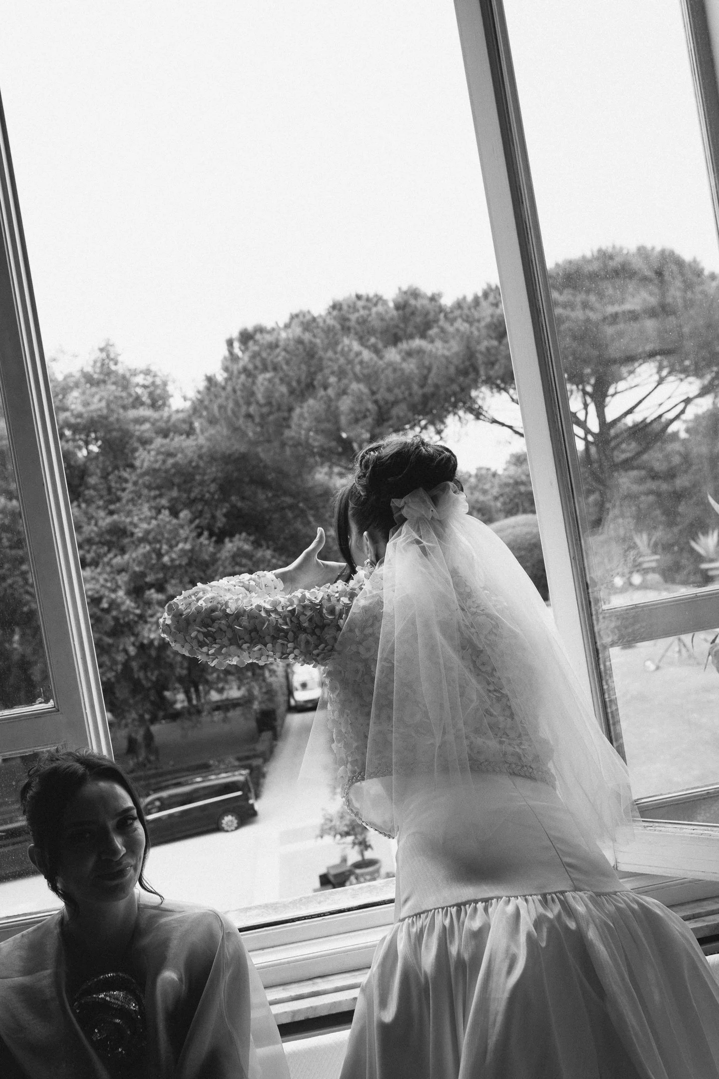 A bride in a wedding dress and veil looking out a large open window with two bridesmaids sitting inside.
