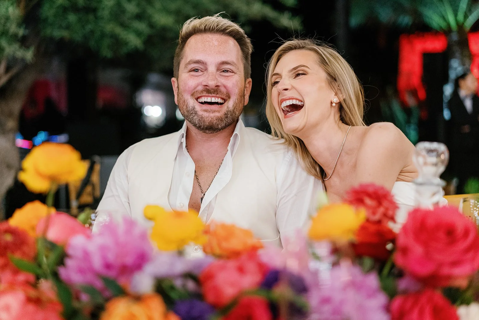 A smiling man and woman sharing a laugh at a dinner or celebration, surrounded by colorful flowers with blurred background.