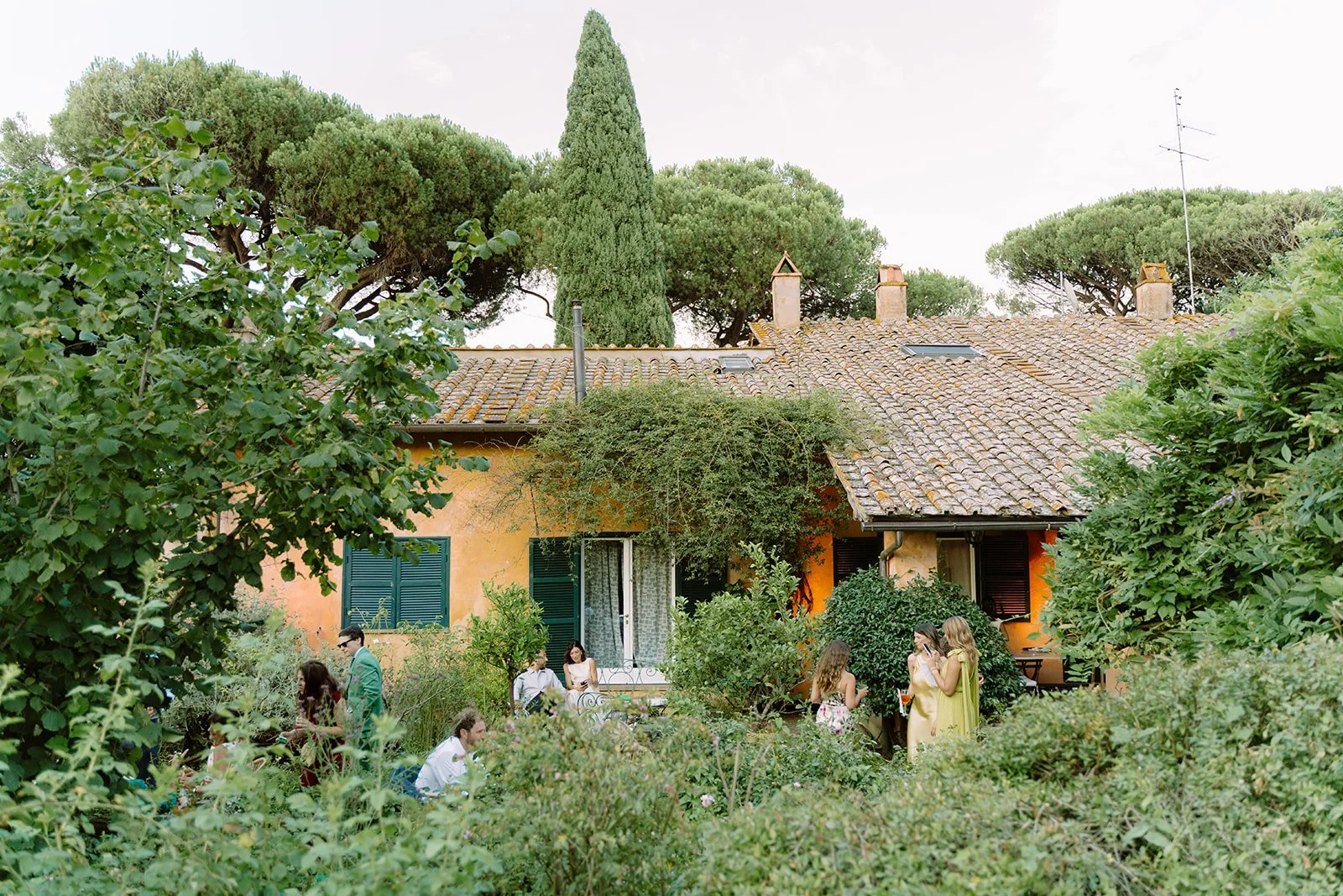 People gathered in a lush garden in front of a rustic orange house with open windows and surrounding green trees and bushes.