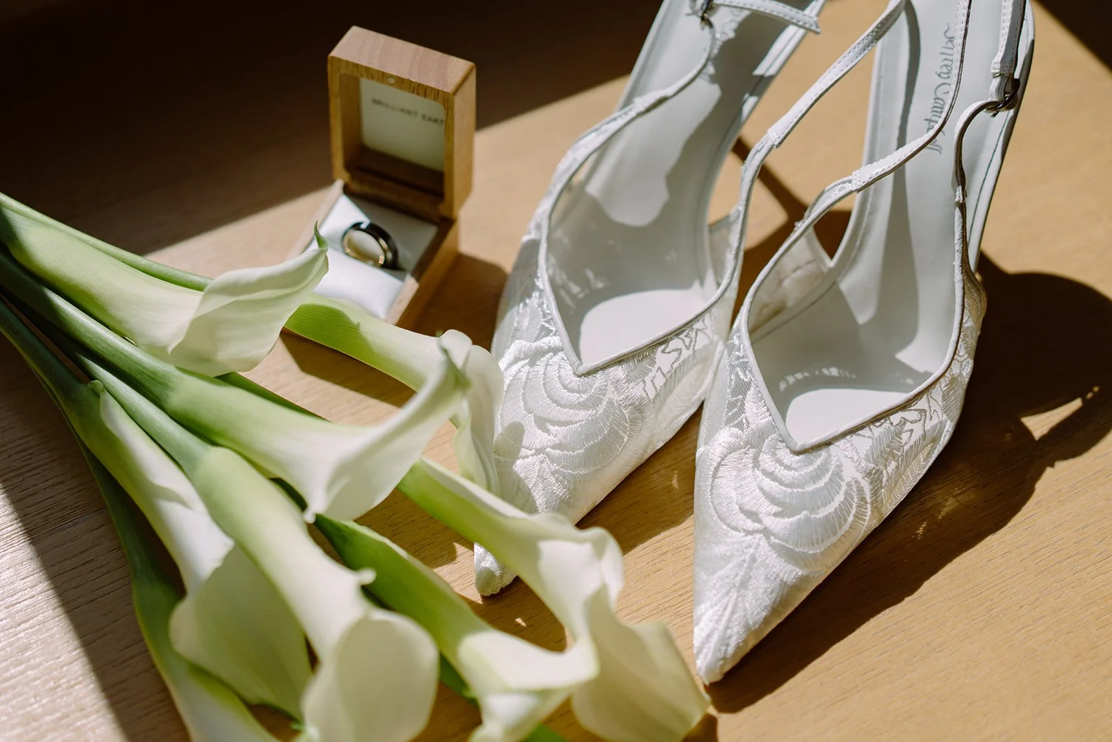A pair of white bridal shoes with floral embroidery, a box with a wedding ring, and a bouquet of white calla lilies on a wooden surface.